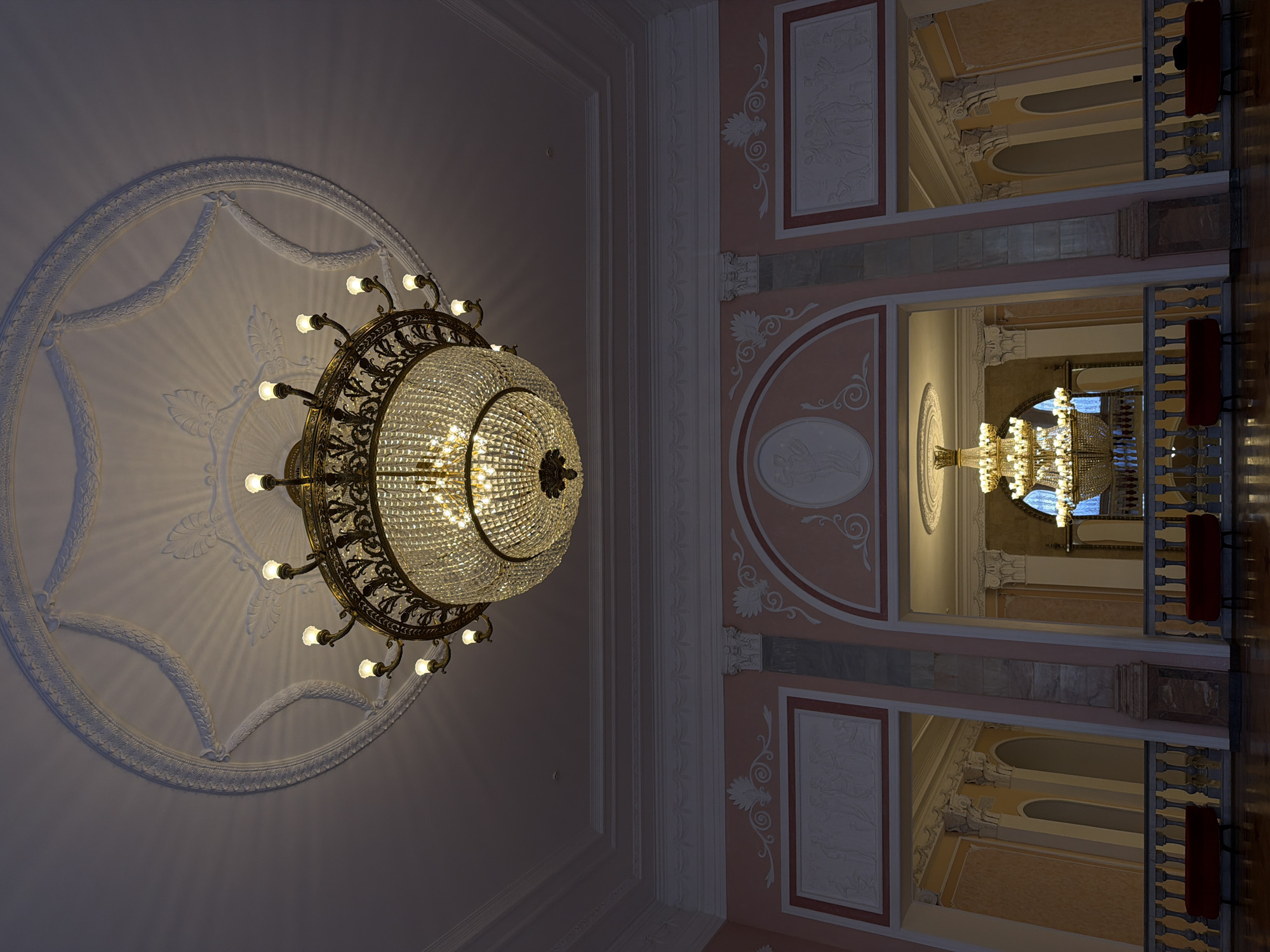 This media file shows an ornate chandelier hanging from a decorative ceiling in what appears to be a grand interior space, possibly a theater or concert hall, given the architectural style and the presence of seating visible in the reflections. The chandelier is a focal point, made of intricate metalwork and numerous glass crystals that catch and refract the light from the bulbs. It hangs within a circular, relief-decorated ceiling medallion.
The surrounding walls are decorated with panels featuring relief sculptures and ornate trim in shades of pink and white. In the reflections, one can see a balcony or upper level with a railing and red seating. The overall impression is one of opulence and classical elegance. There is no discernible text, and the lighting suggests an indoor, likely evening setting. The scene is static, with no people or specific events depicted, focusing solely on the architectural and decorative elements of the location. The location context is Ufa, Russia, which hints at the historical or cultural significance of such a building.