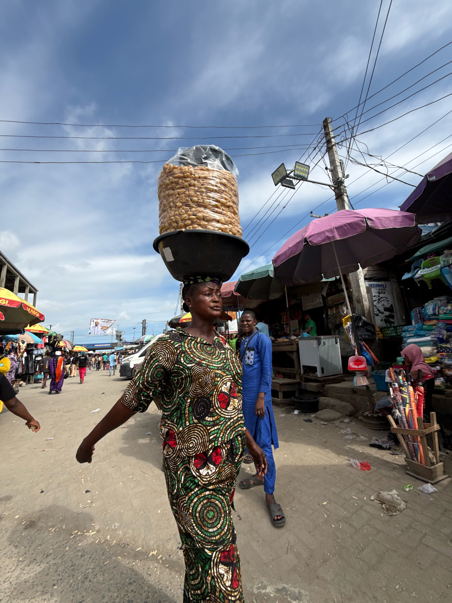 A medium shot captures a bustling marketplace scene in Ado, Nigeria, under a cloudy sky. The central figure is a woman balancing a large, round container overflowing with packaged snacks on her head. She wears a vibrantly patterned dress in shades of green, red, and white with a geometric design. Her head is wrapped in a matching fabric. To her right, a man in a blue traditional outfit walks past, his left hand resting on his hip. In the background, market stalls with colorful umbrellas and awnings are visible, displaying various goods. Several other people are scattered throughout the scene, engaged in shopping or walking. The ground is paved and appears somewhat dusty, with some scattered debris. Utility poles with multiple wires crisscross the sky, indicating an urban setting.