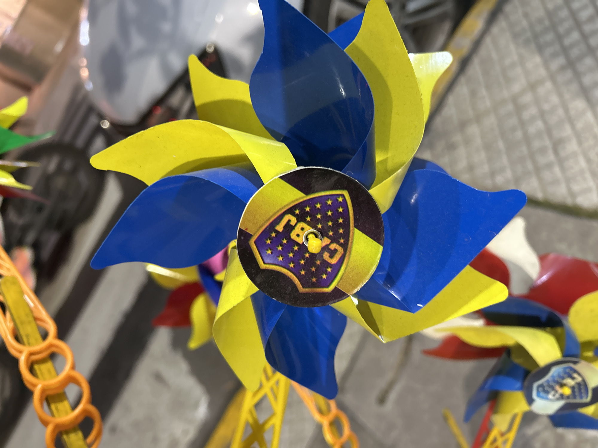 The image shows a blue and yellow pinwheel, with a circular logo in the center featuring stars and the letters "CABJ." This logo likely represents the Club Atlético Boca Juniors, a popular soccer team in Buenos Aires, Argentina. The pinwheel is likely a souvenir or decoration related to the team. The location is probably in Buenos Aires, given the reference to Boca Juniors. Other pinwheels with different colors can be seen in the background. The pinwheel is mounted on a yellow plastic stick with orange circle-shaped segments. The scene appears to be outdoors with a blurry background.