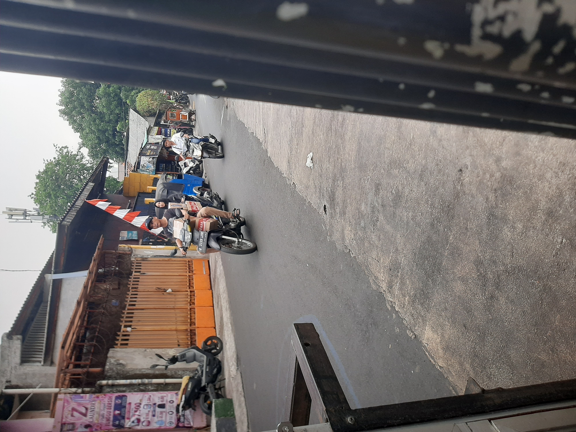 The image depicts a street scene in Central Jakarta, Indonesia, likely a residential or commercial area. The foreground is dominated by a dark, corrugated metal fence, obscuring the left side of the frame. To the left of the fence, a portion of a dark wooden structure, possibly a bench or table, is visible.
The street itself is paved and appears to be wet in places, suggesting recent rain or a damp environment. Several motorbikes are present, some parked and others in motion. On the left side of the street, a dark-colored scooter is parked next to a building with an orange-colored gate. A laundry advertisement banner is attached to the wall of this building.
Further down the street, a group of motorbikes are moving. One rider, a male wearing a dark t-shirt and a baseball cap, is on a motorbike with a white and red flag attached to it. Another motorbike, ridden by a man in a white shirt, is also visible. In the background, several buildings are visible, some with tiled roofs and others with corrugated metal roofs. Trees are interspersed with the buildings, indicating the presence of greenery in the urban landscape. The sky is overcast, suggesting a cloudy day.
