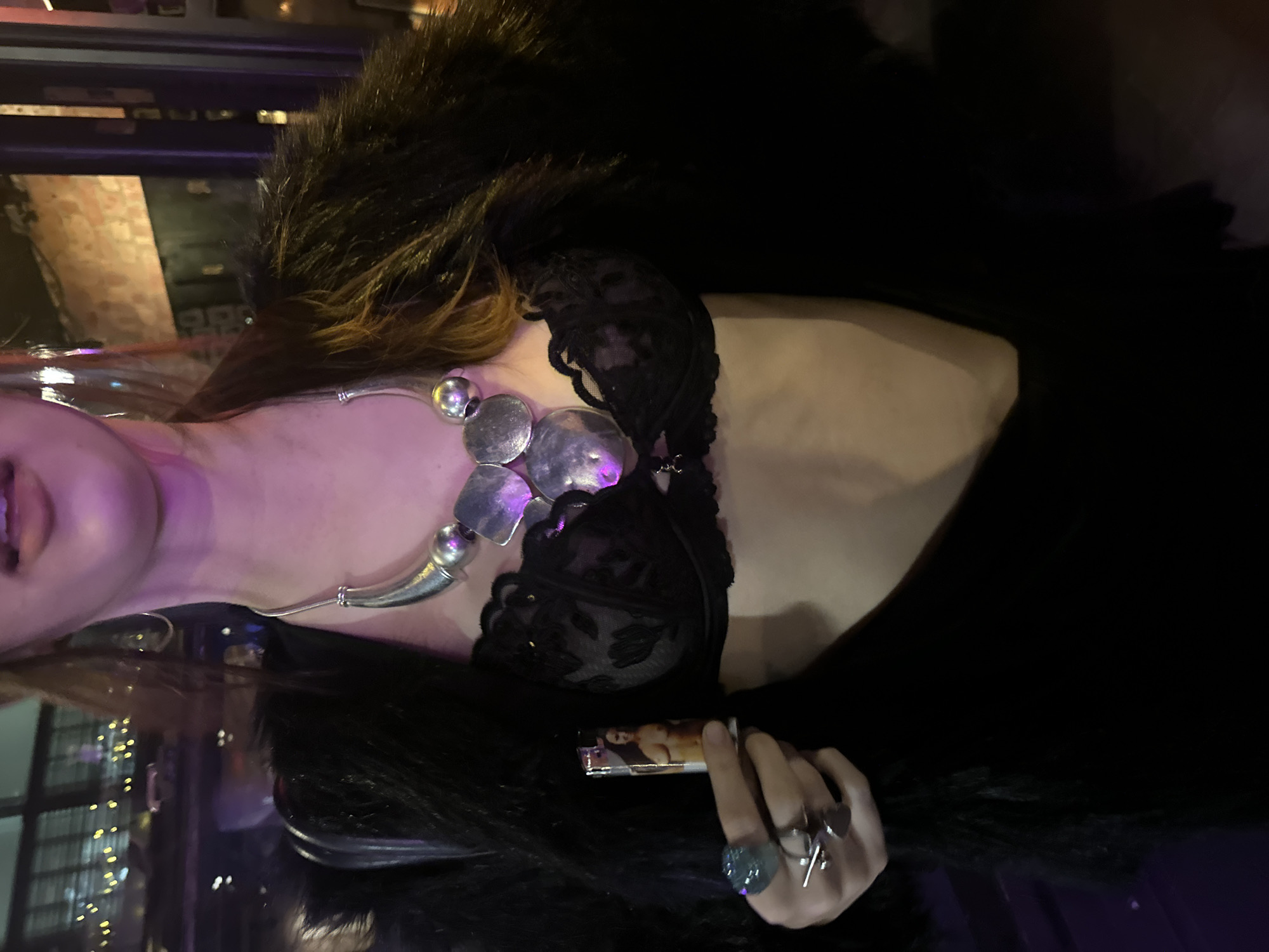 A person with pale skin and pink lips is partially visible in a dimly lit indoor setting. They are wearing a black lace bra, a statement silver necklace with large, irregular pendants, and a dark fur stole or coat. The person is holding a lighter with an image of a nude woman on it. Two rings are visible on their fingers. In the background, there are hints of an urban environment, possibly in London, United Kingdom, suggested by the architectural style and possible presence of brick. String lights are also visible in the background.