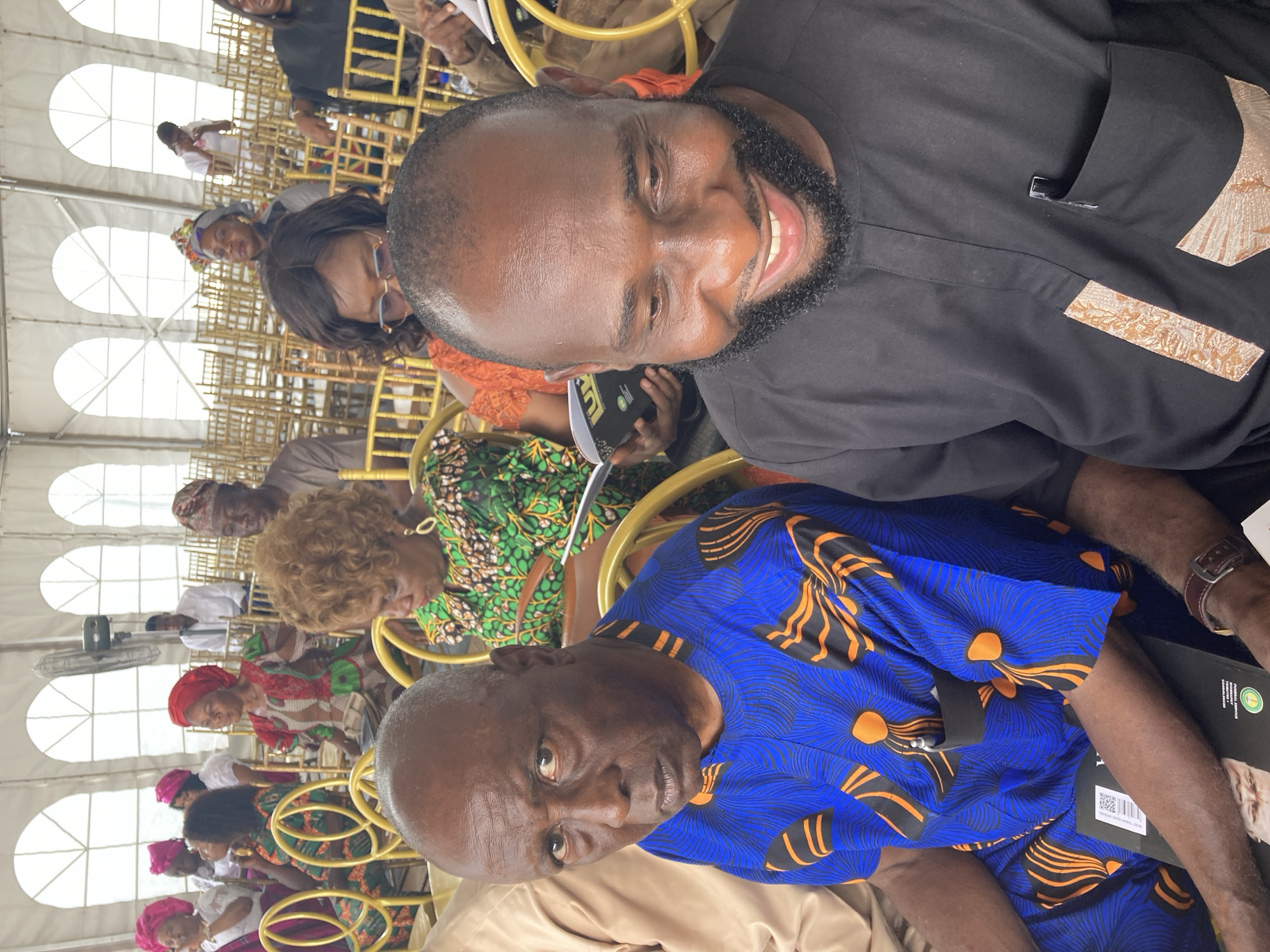 A medium-angle shot depicts two men and a crowd of people, likely at an event in Nnenu, Nigeria. The man closest to the viewer is wearing a blue shirt with orange and black floral patterns. Behind him is a younger man with a beard, wearing a black shirt with patterned gold details. Both men are smiling and looking slightly down, likely at something or someone. The background shows rows of people seated in gold chairs, dressed in various traditional Nigerian attire including head wraps and patterned fabrics. The setting appears to be indoors under a white tent-like structure.