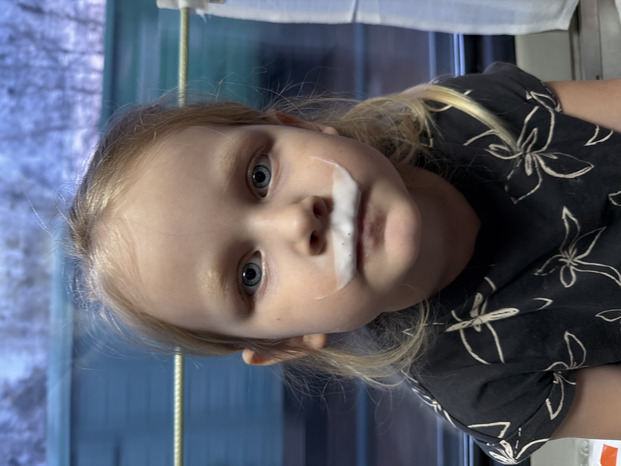 A close-up, candid portrait features a young child with fair skin, light blonde hair, and prominent blue eyes, looking directly at the camera. A distinct "milk mustache" or white foam is visible above the child's upper lip, indicating recent consumption of a beverage. The child wears a dark-colored t-shirt adorned with a repeating pattern of white floral designs. Their head is slightly tilted, and their expression appears calm and engaging.
The background, viewed through a window, is softly blurred, creating a shallow depth of field. This exterior scene reveals what appears to be a wintry or early spring landscape with light-colored, bare trees or bushes against a bright, possibly overcast sky, suggesting it is daytime. Interior elements like a window frame, a white curtain along the top edge, and a vertical light-colored support bar are also discernible, suggesting the child is indoors, potentially within a vehicle such as a train or bus. The overall mood is spontaneous and playful. No visible text is present in the image. The location context for this scene is Michurinsk, Russia.