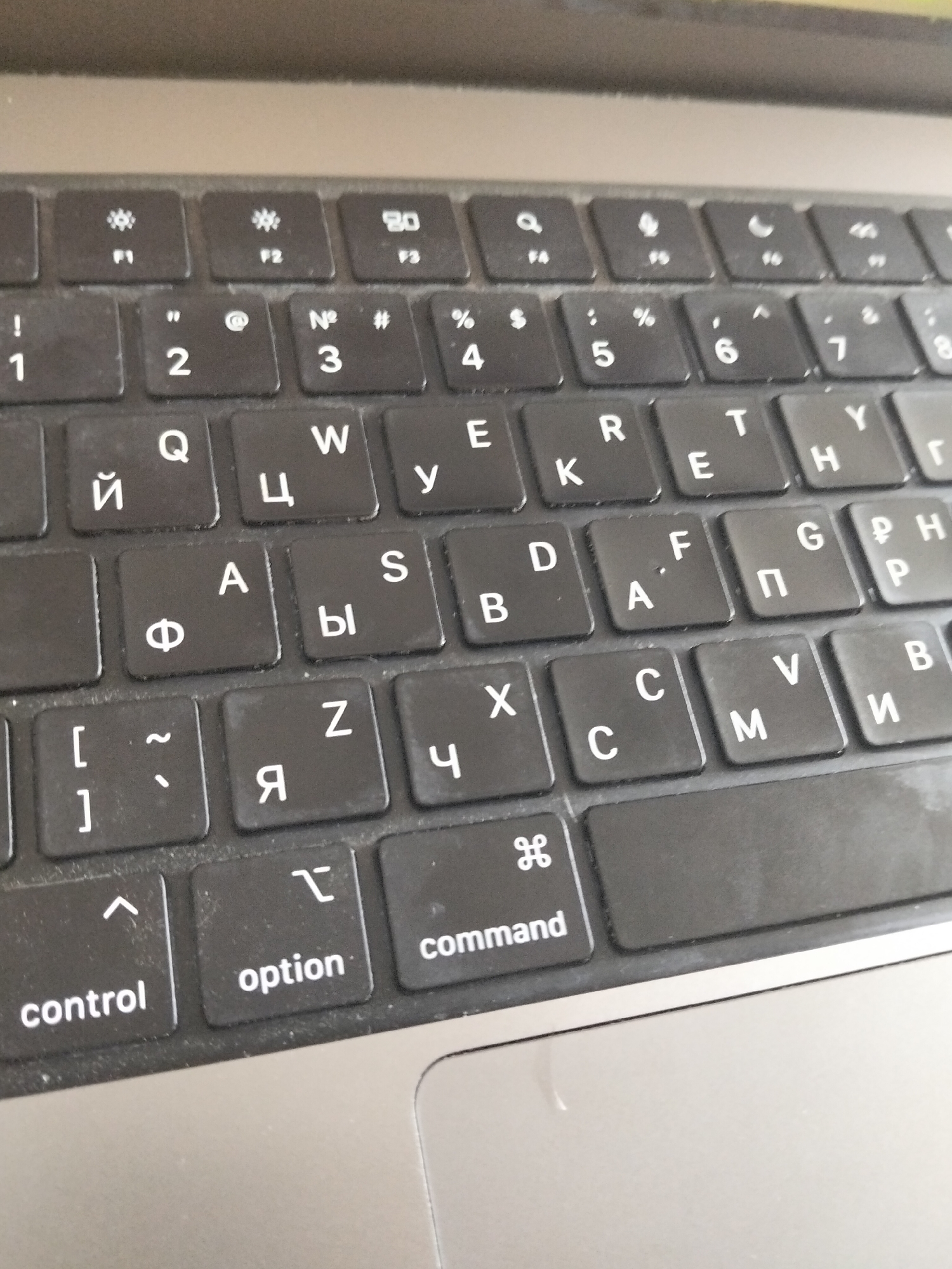 The image shows a close-up, overhead view of a black laptop keyboard. The keyboard is angled slightly, with the top edge of the laptop visible at the very top of the frame. The keys are mostly black with white lettering, and some keys also have a symbol above the letter. Some keys display Cyrillic letters, suggesting it's a Russian keyboard layout. Specific keys visible include numbers 1-8, function keys F1-F7, and letters like Q, W, E, R, T, Y, A, S, D, F, G, H, Z, X, C, V, B, M, and Cyrillic equivalents such as Й, Ц, У, К, Е, Н, Я, Ф, Ы, В, A, П, М, И. Additionally, modifier keys like "control", "option", and "command" are visible on the bottom row. The keyboard appears to be slightly dusty, with a few faint smudges on the keys and surrounding surface. The lighting is overhead and somewhat diffused. There are no people or other objects visible in the frame. The image is a static shot of a technological device.