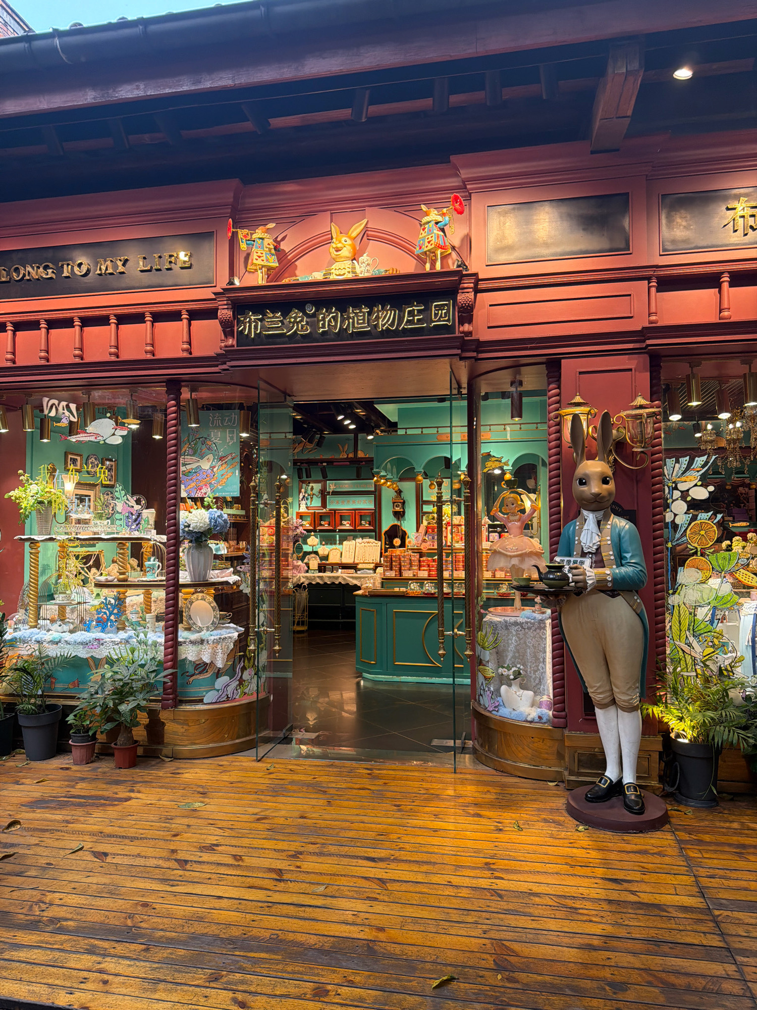 The image shows the exterior of a shop in Chancheng District, China. The shop has a red facade with gold lettering that reads "SONG TO MY LIFE" and Chinese characters above the entrance. A large statue of a rabbit dressed in a blue jacket and tan breeches, holding a tea tray, stands to the right of the entrance. The shop windows display various items, including decorative plants, ceramics, and figurines, against a teal-colored interior. The shop floor is made of weathered wooden planks. The overall scene depicts a whimsical and decorative storefront.