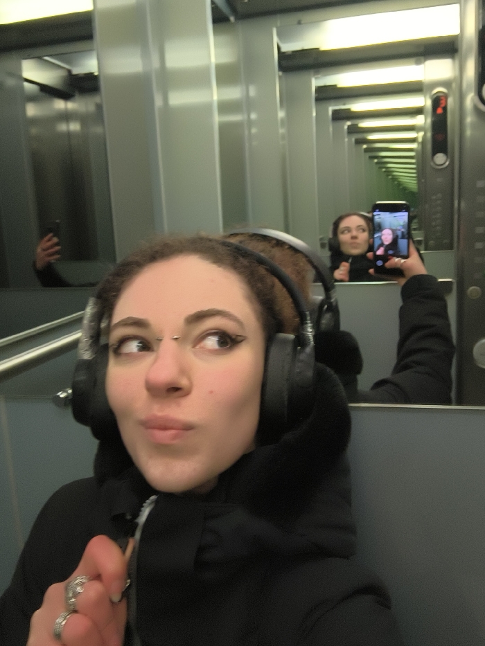 A young woman with curly brown hair, wearing black over-ear headphones and a dark hooded jacket, is captured in a selfie taken in an elevator. She is positioned in the foreground, her face turned slightly to the right with a playful, slightly puckered expression and a side-long glance. She has prominent black winged eyeliner and a silver bridge piercing above her nose. On her left hand, which is partially visible near her chest, she wears several rings.
The setting is an elevator with highly reflective, possibly mirrored, walls. This creates a striking visual effect of infinite reflections, showing multiple receding images of the woman and the elevator interior. In the reflection directly behind her, the woman is seen holding up a black smartphone to take the picture, her face focused on the phone screen. Another reflection further into the distance shows her hand holding the phone again. On the right side of the main reflection, part of the elevator's control panel is visible, displaying the number "2", likely indicating a floor. The environment is modern and metallic.
