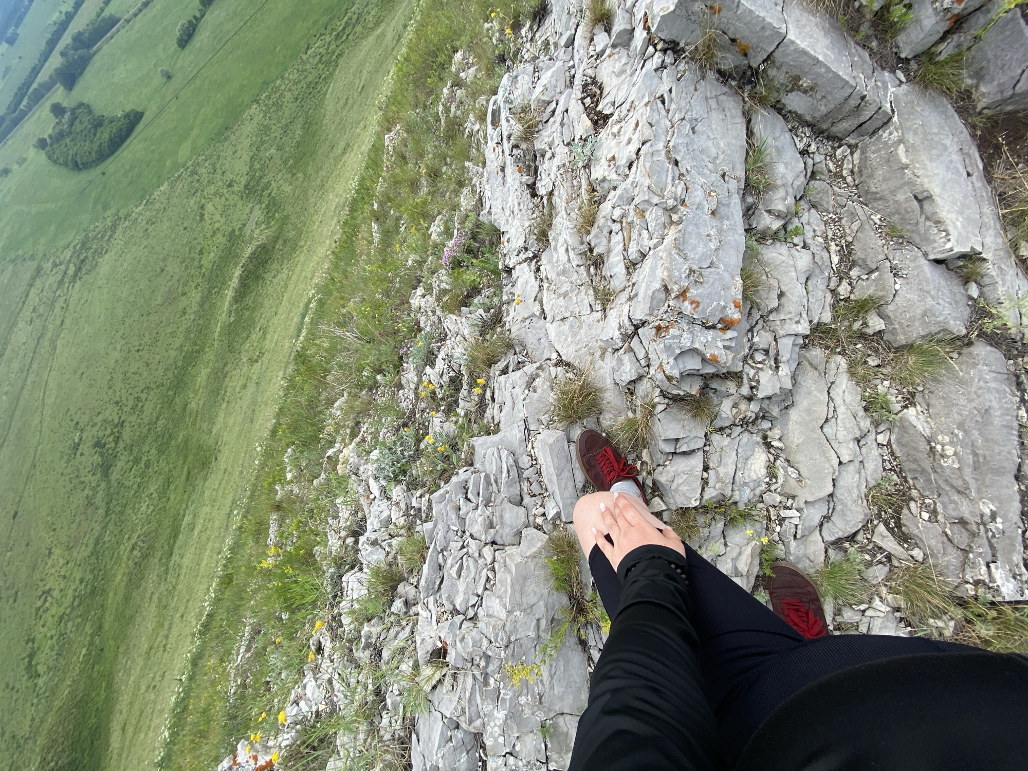 The image shows a person's legs and feet sitting on a rocky outcrop overlooking a steep, grassy slope. The person is wearing black pants and red shoes. The terrain is mountainous and covered in green grass and scattered wildflowers. The sky is not visible in the frame. The location is stated to be in the city of Verkhnyaya Chernorechka, Russia.