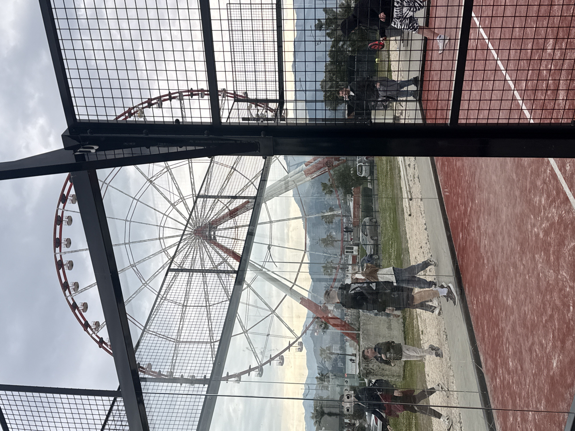 The image, taken in Batumi, Georgia, captures a view through the glass and mesh enclosure of what appears to be a paddle tennis court. Reflected in the glass are people walking by, as well as a Ferris wheel. A red tennis court is visible with people playing. Beyond the people and amusement ride, distant mountains are visible under an overcast sky.