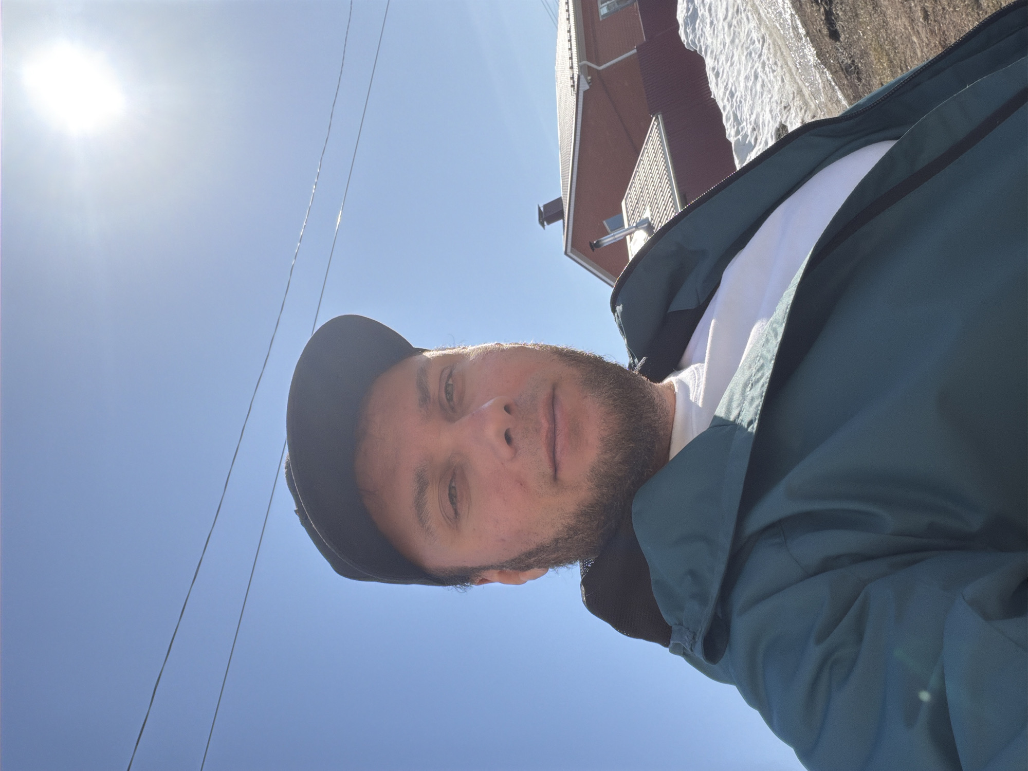 The image is a selfie of a man wearing a dark baseball cap, a white t-shirt, and a green jacket. He has a beard and is looking directly at the camera.
The background includes a bright blue sky with the sun shining and a few power lines. A corner of a building with red siding and snow on the ground is visible.
The photo was taken outdoors in Chaikovsky, Russia.