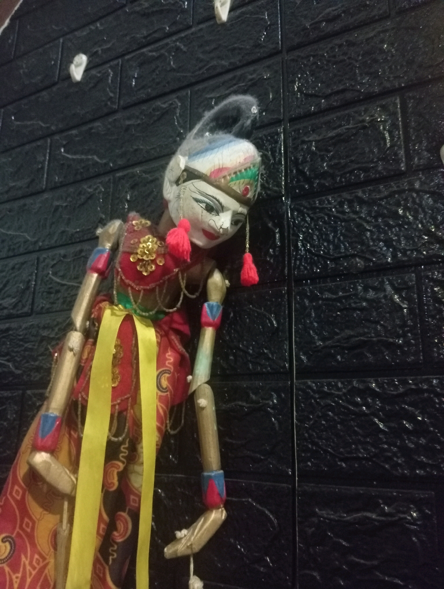 A full-body shot shows a detailed, traditional Javanese Wayang Golek puppet with a white head, dark eyes, and red lips, leaning against a textured black brick wall. The puppet is adorned with a red and gold costume featuring intricate patterns, a yellow sash, and red tassels. Its wooden limbs are visible, with colorful detailing on the forearms. The background is a dark, slightly glossy, faux-brick wall with subtle highlights from overhead lighting. The image appears to have been taken indoors, likely in a home or cultural setting in Garut, Indonesia.