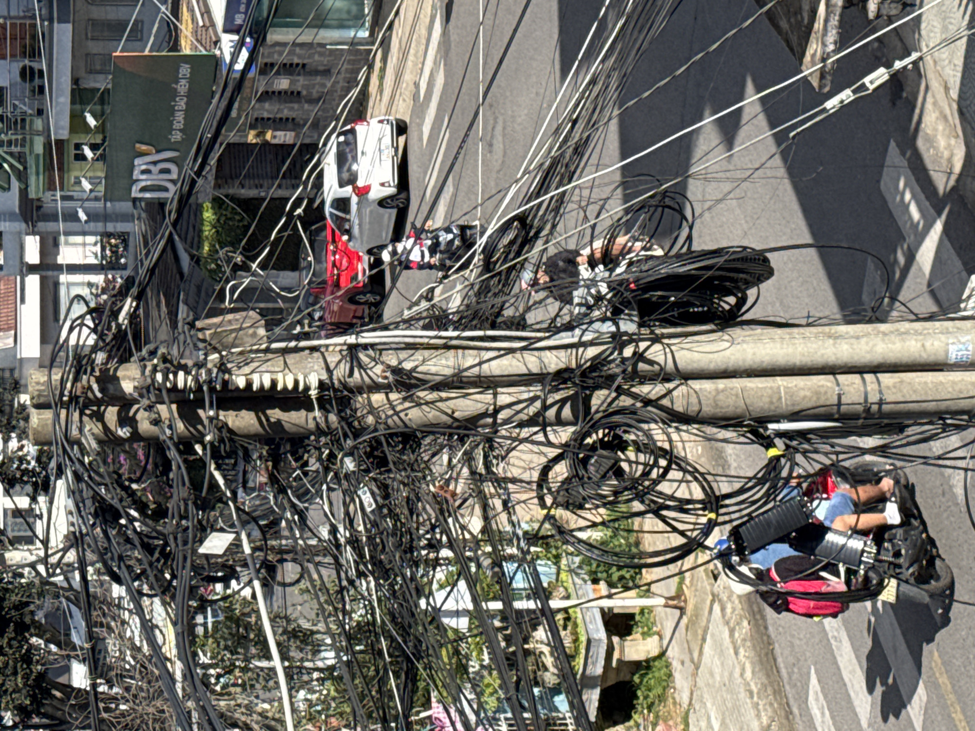 The image depicts a dense urban street scene in Da Lat, Vietnam, characterized by a substantial tangle of black electrical and telecommunication wires hanging precariously from a concrete utility pole. A red car and a white car are visible on the asphalt road, with a pedestrian crossing marked by white stripes. Two individuals on motorcycles are also present. One motorcycle, carrying a driver and a passenger with a red backpack, is positioned in the foreground, partially obscured by the utility pole and its wires. Another motorcycle, with a single rider, is further down the street. Buildings with visible signage, including "DBV TẬP ĐOÀN BẢO HIỂM DBV," line the street. The environment suggests a busy, developed urban area with active transportation.