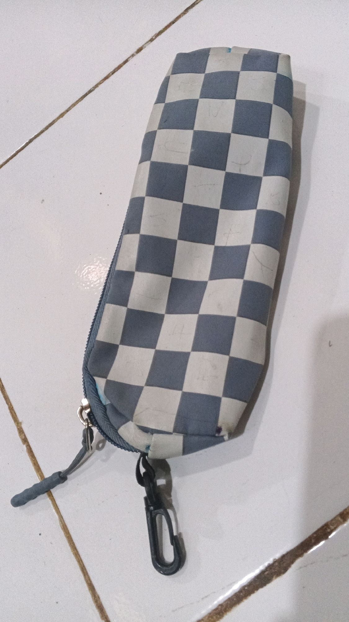 The image shows a checkered pencil case on a tiled floor. The pencil case is a light gray and white checkerboard pattern. It has a zipper closure, a small gray zipper pull, and a black carabiner clip attached. The tiled floor appears to be white with thin, brown grout lines. The image is taken from a top-down perspective. The location in Winong, Indonesia is not ascertainable from the image.