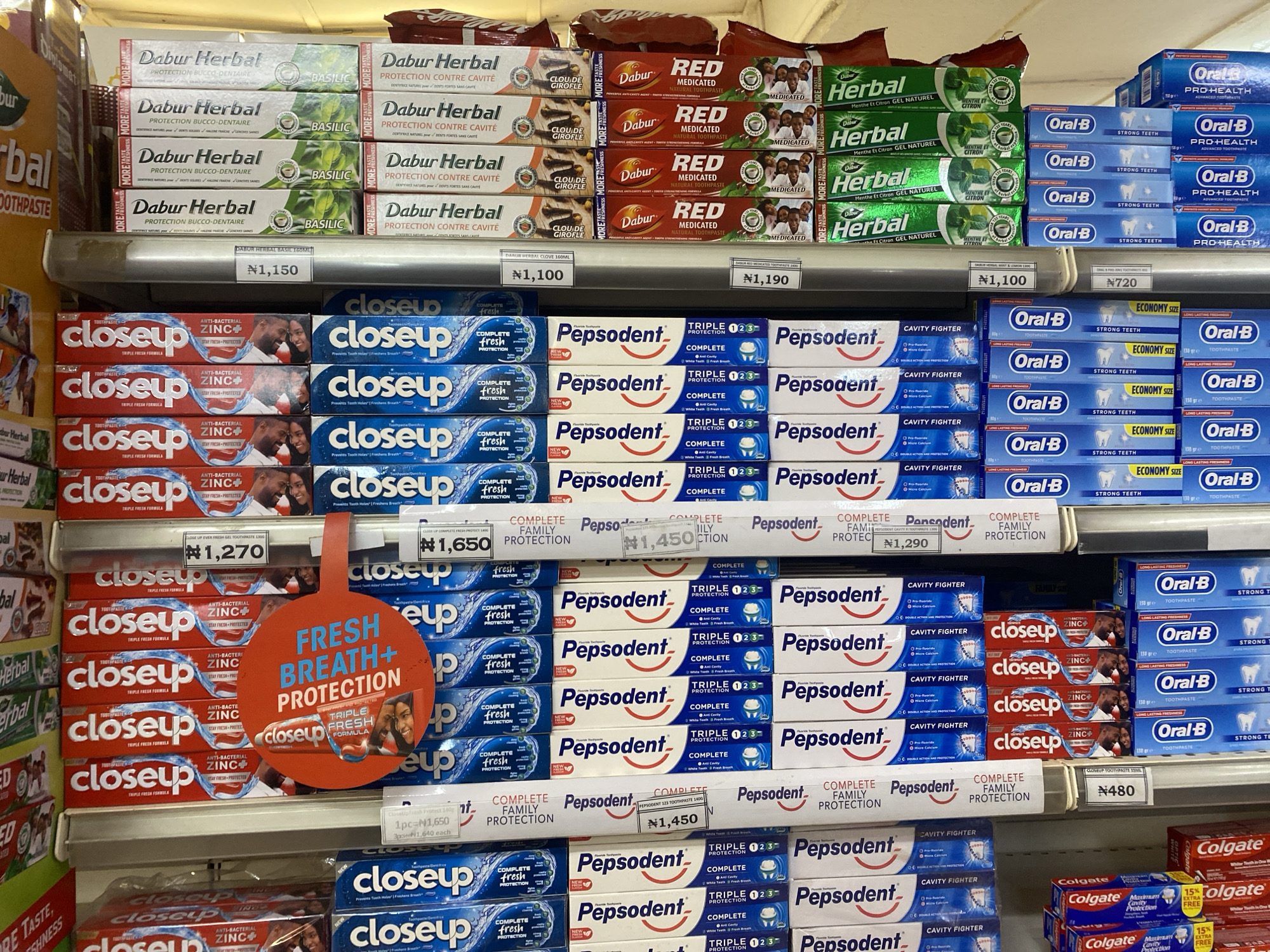 The image shows a retail shelf in a store, likely a supermarket or pharmacy, in Ikorodu, Nigeria. The shelves are stocked with various brands of toothpaste. Prominently displayed are rows of Dabur Herbal, Close Up, Pepsodent, and Oral-B toothpastes. Several items have price tags attached, indicating individual prices for some products and bulk pricing for others. A large orange sign with "FRESH BREATH+ PROTECTION" written on it hangs from the shelf, promoting a Close Up toothpaste product. The overall environment appears to be a well-organized commercial display of consumer goods.