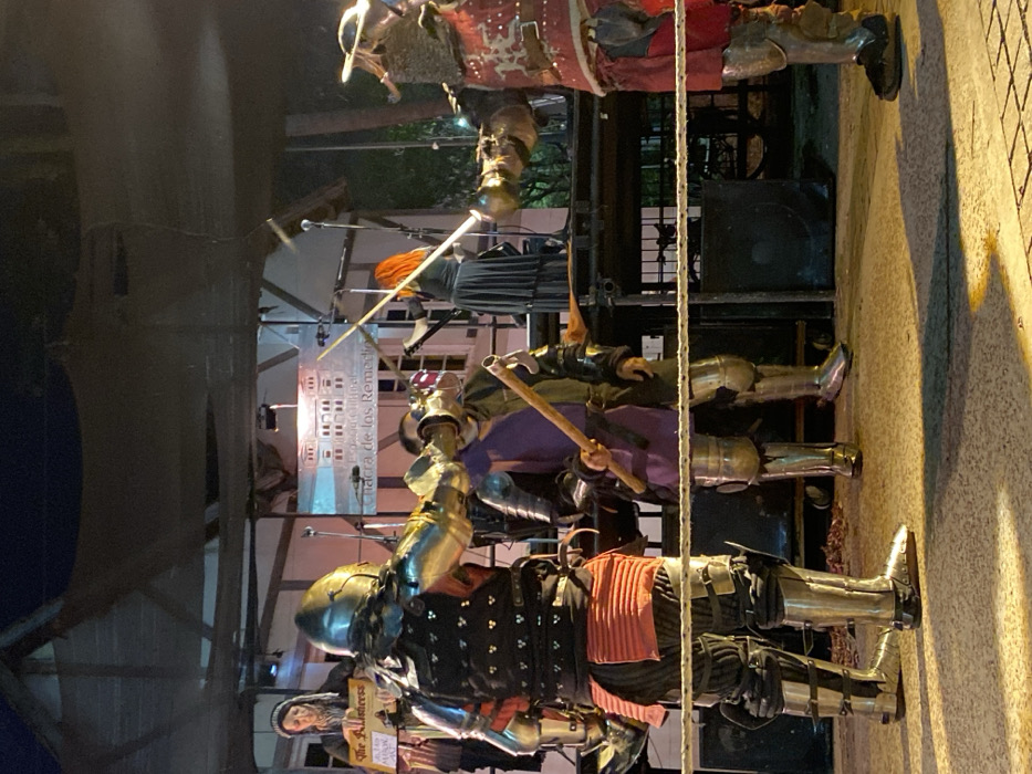 This image depicts a historical reenactment or performance taking place at night at the Espacio Cultural Chacra de los Remedios within Parque Avellaneda, Buenos Aires, Argentina.
In the foreground, three individuals in full plate armor are engaged in a simulated combat. The leftmost armored participant, wearing a dark cuirass over red and black segmented undergarments, extends a gauntleted right hand forward as if striking. The central armored participant, dressed in a purple tunic over shining plate armor, holds a wooden hafted weapon vertically. The rightmost armored participant, wearing red and white segmented armor, extends a long polearm towards the center. A white rope barrier runs horizontally across the lower middle ground.
On a raised stage in the background, two additional individuals are present. On the left side of the stage, a woman in a dark medieval-style dress and head covering holds a sign reading 'The Alchemist.' To her right, another woman with long red hair, dressed in a dark gown, wields a long, slender sword-like object. A banner behind them prominently displays 'Espacio Cultural Chacra de los Remedios' alongside a stylized building illustration. The outdoor scene is illuminated by artificial stage lighting, and two large sound speakers are positioned beneath the stage structure. The ground appears to be paved.