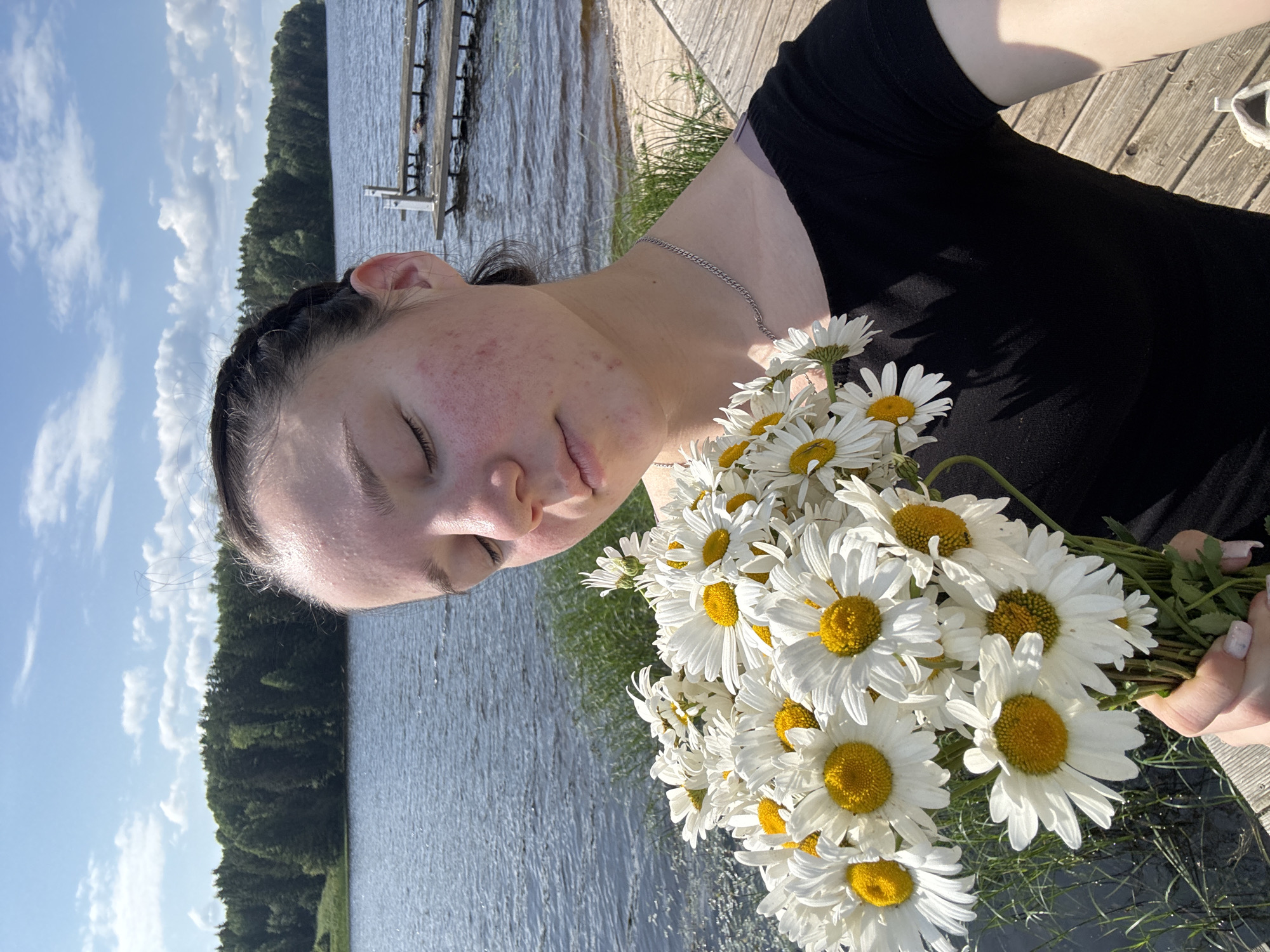 This is a vertical, close-up, outdoor, selfie-style shot of a young person holding a bouquet of daisies. The subject is positioned on the right side of the frame, their face partially visible with eyes closed, head tilted back slightly. Their skin shows some redness, particularly on the cheeks and forehead. They are wearing a black t-shirt and a silver chain necklace. The bouquet of daisies is held in the lower right corner of the frame, obscuring part of the subject's chest and arm.
The background features a body of water, likely a lake, with a treeline on the far side. A wooden dock or pier is visible in the upper right portion of the image, with some wooden structures extending into the water. The sky above is blue with scattered white clouds. The lighting suggests it is daytime, likely late afternoon or early evening, given the warm glow and shadows. The overall mood appears peaceful and serene, perhaps evoking a sense of enjoying nature. The location is identified as Pozdyshki, Russia, which aligns with the naturalistic scenery. There is no visible text in the image.