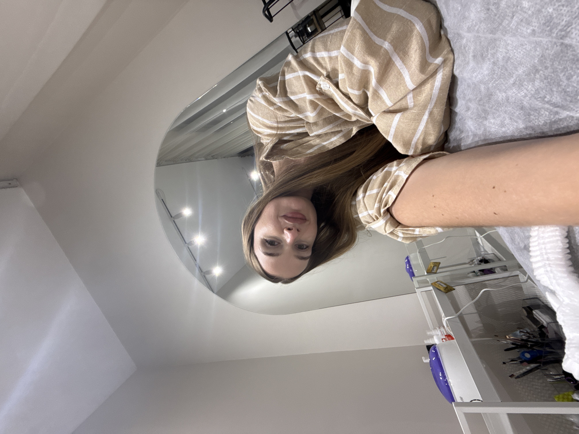 The image, taken in Budva, Montenegro, shows a woman taking a selfie reflected in a large, curved mirror. The woman, appearing to be in her 20s or 30s, is lying on a light gray, textured surface, possibly a treatment bed or table. She has long brown hair and is wearing a beige shirt with white stripes. Her upper body is visible, with her face looking directly towards the camera in the reflection.
The setting appears to be a professional environment, likely a beauty salon or spa. Bright ceiling lights are visible and reflected prominently in the mirror, illuminating the space. To the right of the frame, a multi-tiered white utility cart is partially visible. On its top shelf, a white and purple device, possibly related to beauty treatments, can be seen. Below it, various tools, brushes, and containers are arranged on the lower shelves. The walls and ceiling are white, suggesting a clean and modern aesthetic. The image is captured from a slightly upward angle, with the woman's reflection dominating the upper half of the frame. No clearly legible text is visible in the image.