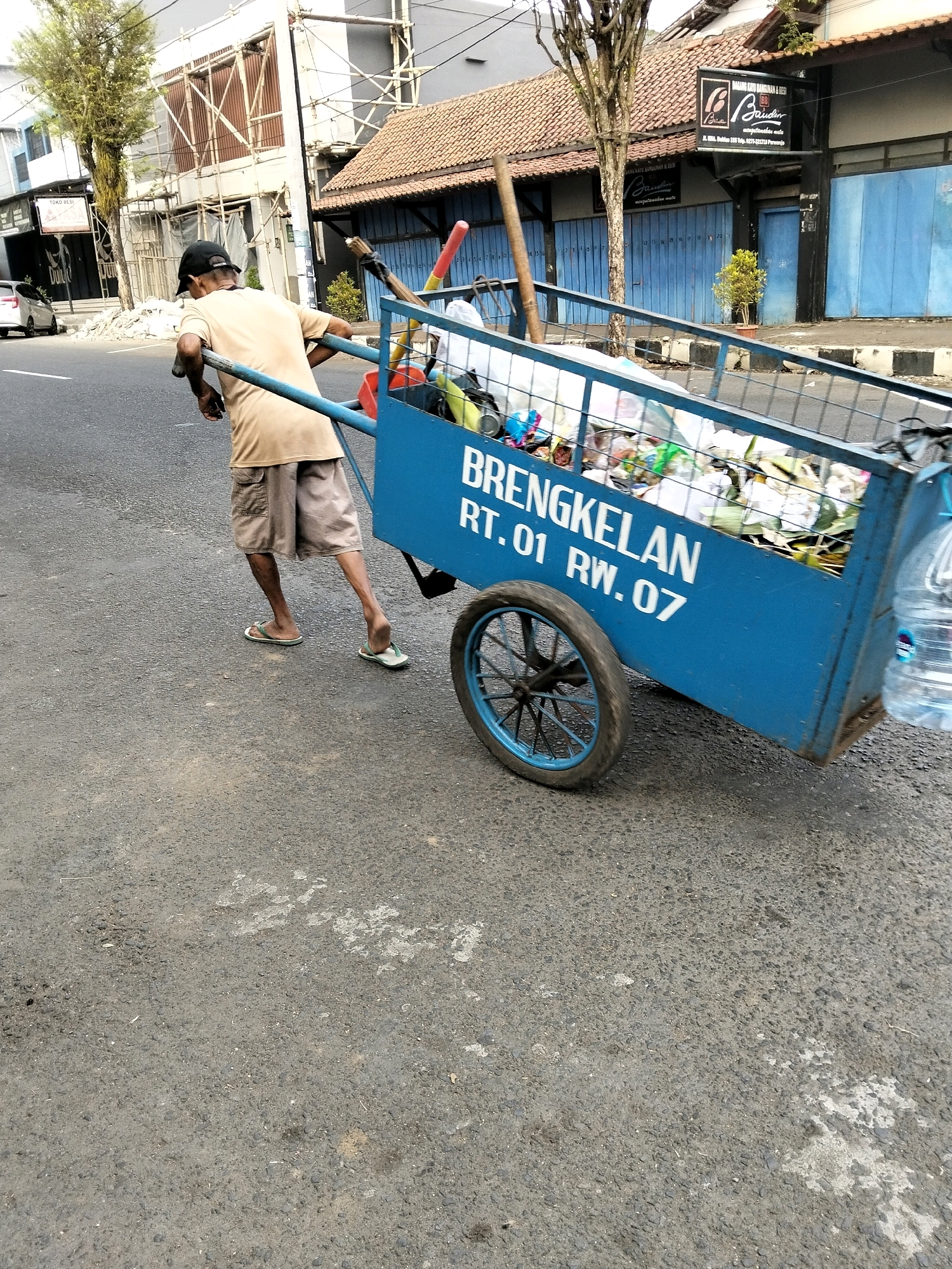 A man wearing a beige t-shirt, khaki shorts, a black baseball cap, and flip-flops pushes a blue cart filled with trash down an asphalt street in Purworejo, Indonesia. The cart has "BRENGKELAN RT.01 RW.07" printed on its side. The man is viewed from the back, with his left arm resting on the cart's handle. Behind the cart, a building with a tiled roof and blue shutters is visible. A tree with bare branches stands in front of the building. The street is lined with what appears to be a sidewalk or curb. A white car is parked on the left side of the street. Construction scaffolding is present on another building in the background. The overall scene suggests an outdoor urban environment.