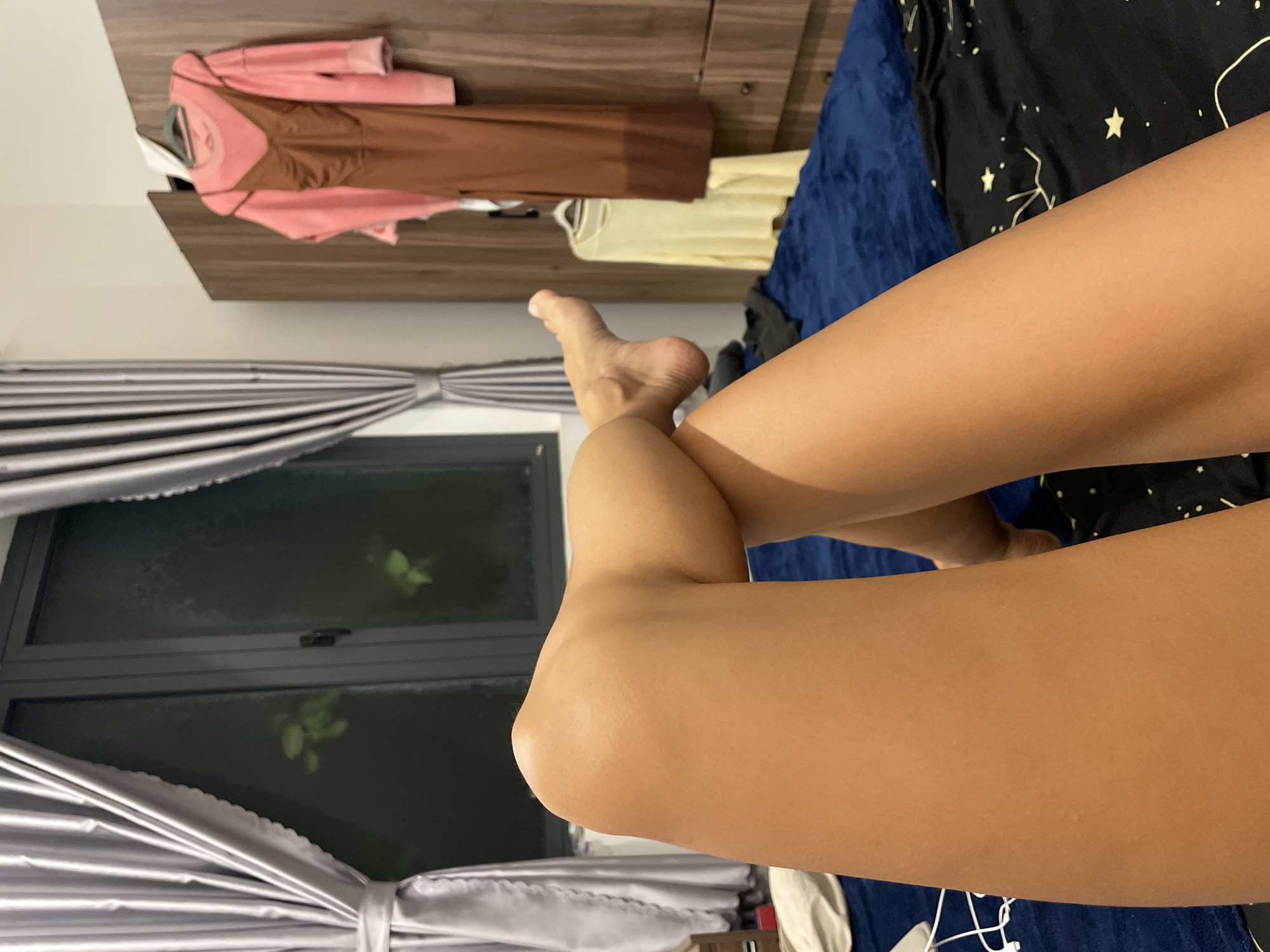 The image captures a first-person perspective of a person's legs and feet, suggesting a moment of relaxation indoors. The person's lightly tanned legs are crossed, with one foot partially visible, resting on what appears to be a bed.
In the background, to the left, a window is partially framed by grey, pleated curtains that are tied back. The view outside the window is dark, with faint green foliage visible, indicating it is nighttime or evening. Above the window area, a wooden wardrobe or shelving unit is visible, from which several items of clothing are hanging: a pink long-sleeved top, a brown slip dress, and a light yellow top.
The bed on which the person is resting is covered by a dark blue textured blanket on one side and a dark fabric patterned with white stars and abstract lines on the other. White earphones are visible near the bottom right edge of the frame. The overall setting is an indoor space, likely a bedroom, conveying a sense of leisure in the evening or night in Mũi Né, Vietnam. No visible text is present in the image.