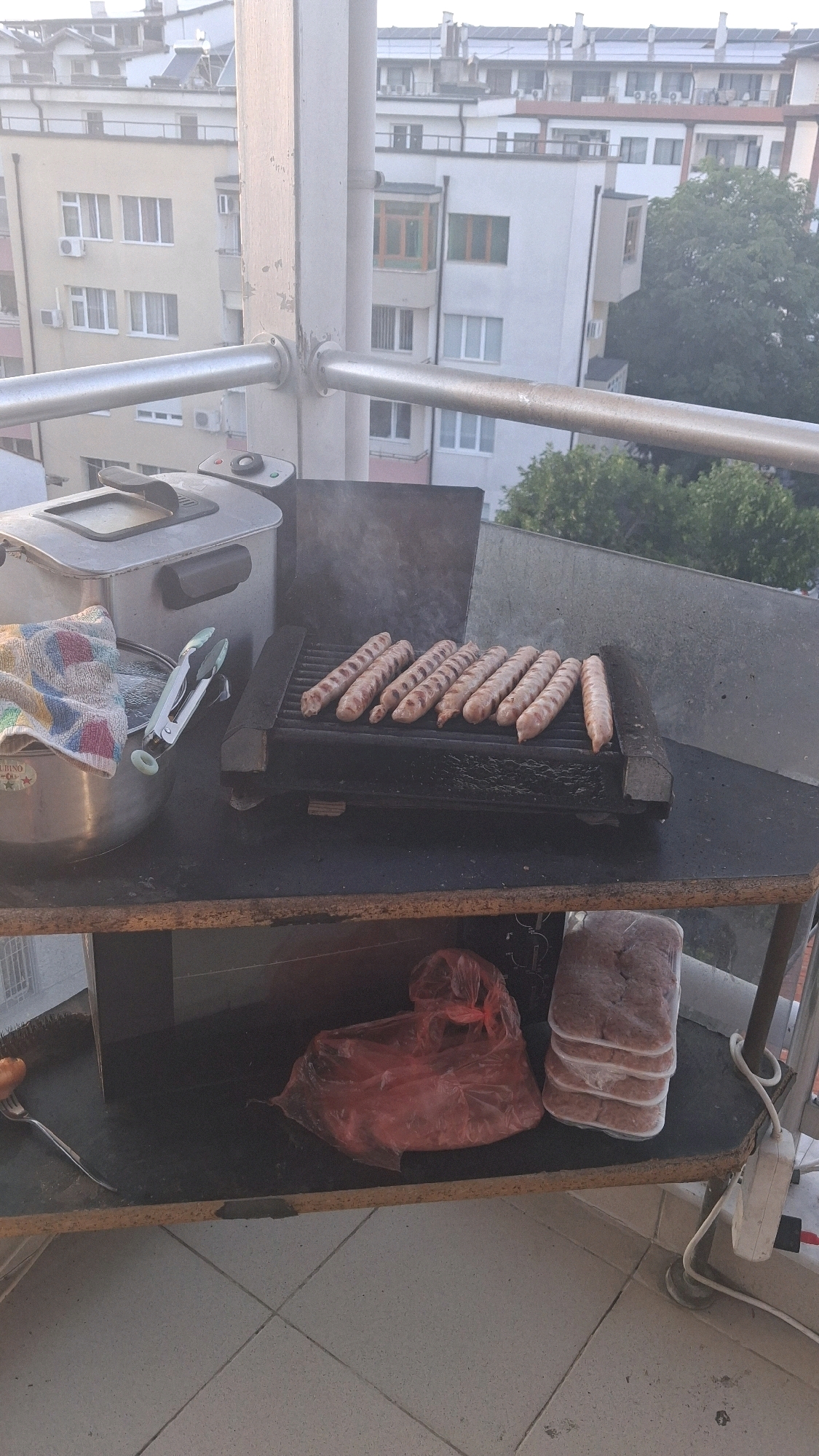 The image captures an outdoor scene on a balcony, likely in the city of Gotse Delchev, Bulgaria. In the foreground, a black electric grill is set up on a dark, worn table. Several sausages are actively cooking on the grill, producing visible smoke. To the left of the grill, a stainless steel bowl is present, containing water and a colorful towel, along with a pair of kitchen tongs. Behind these items, a silver deep fryer with its lid closed is visible. The table itself appears to be made of particleboard with visible wear and tear. Below the table, a red plastic bag and a stack of frozen food packages are stored. The background reveals apartment buildings with various architectural styles, common in urban Bulgarian settings, and lush green trees. The overall impression is a casual outdoor cooking setup, possibly for a meal preparation on a warm day.