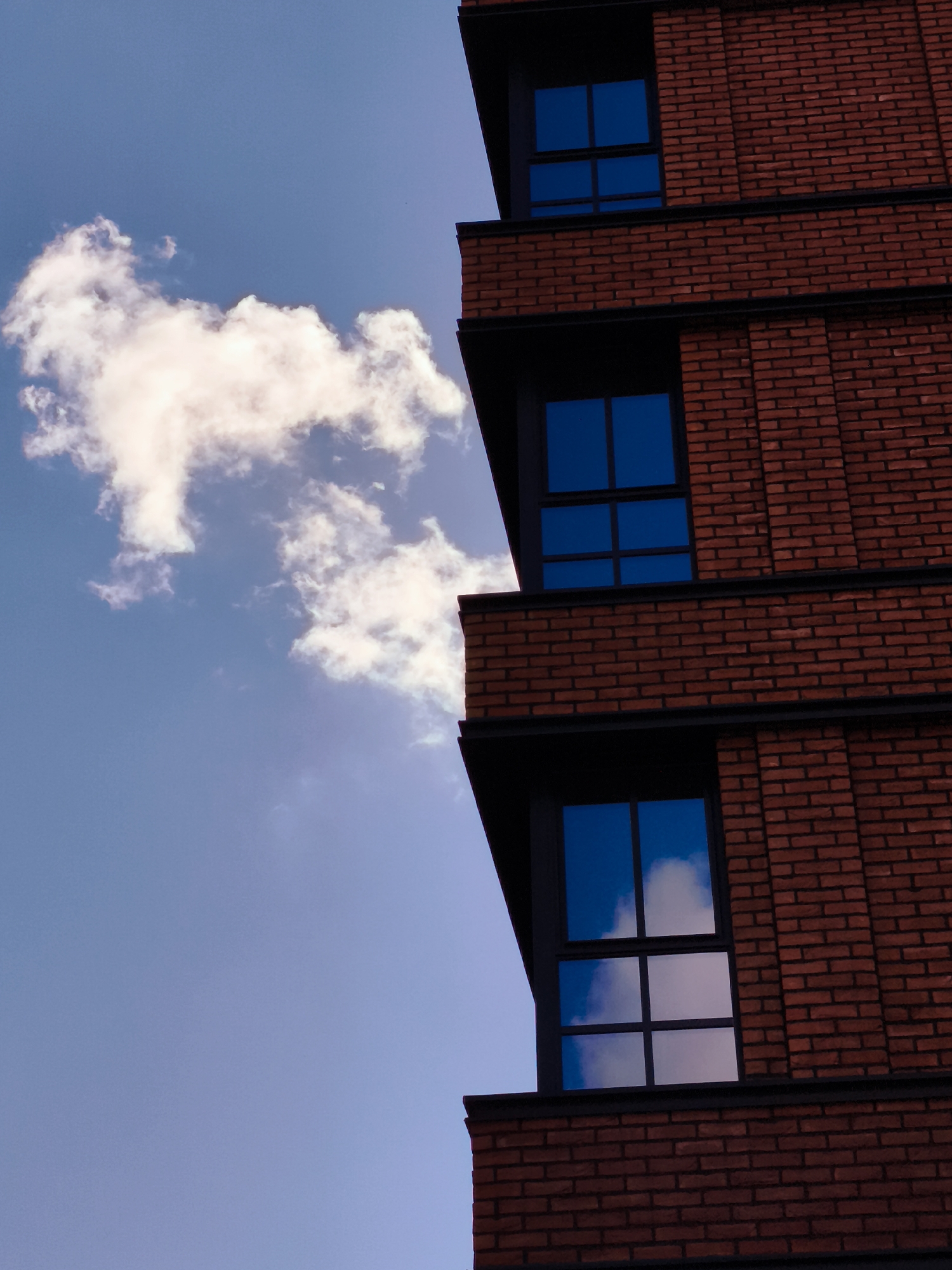 The image shows a close-up, low-angle shot of a modern brick building with blue-tinted windows against a clear sky with a fluffy white cloud. The building's exterior is constructed of dark red bricks laid in a stretcher bond pattern, with dark grey horizontal bands separating the levels. Three windows are visible, each divided into six panes. The sky is a bright, clear blue, with a single large white cloud positioned in the upper left quadrant of the frame. The lower right window reflects the blue sky and parts of the white cloud. The composition emphasizes the geometric lines of the building against the organic form of the cloud and the vastness of the sky. The scene appears to be an exterior view of an urban environment, with no people or specific identifying landmarks visible. The location is identified as Gdańsk, Poland.