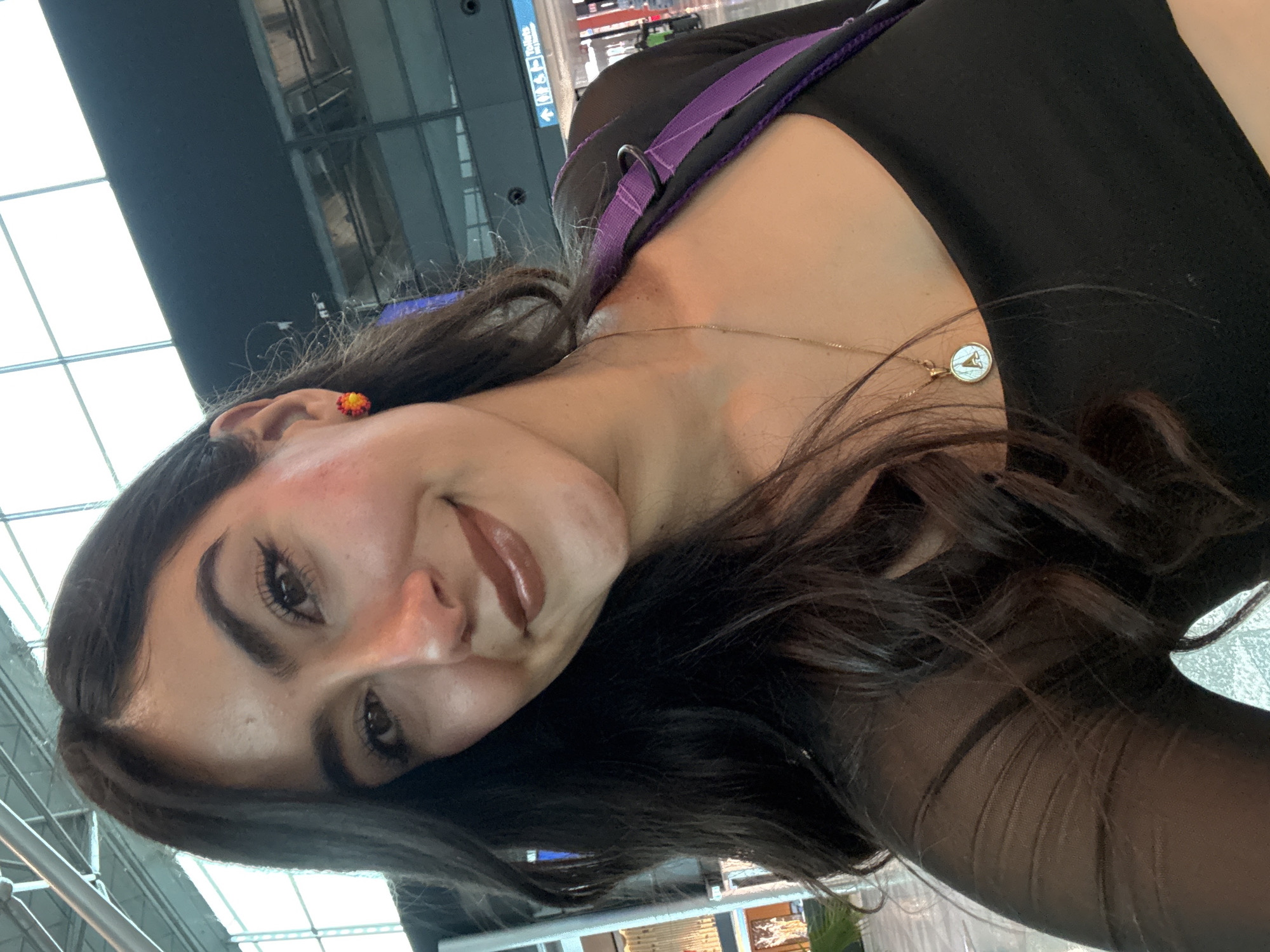 A young woman with long, dark, wavy hair and brown eyes is pictured from the chest up, smiling and looking at the camera. She has on light makeup with noticeable eyeliner and lipstick. She is wearing a black top with sheer sleeves and a gold necklace with a round pendant. A purple strap of a bag is visible over her shoulder. She is in an indoor location with a modern ceiling structure and glass partitions, possibly an airport terminal. The background shows signs with arrows and the word "Toilets" in English and what appears to be Finnish. This scene is located at Helsinki Airport in Vantaa, Finland.