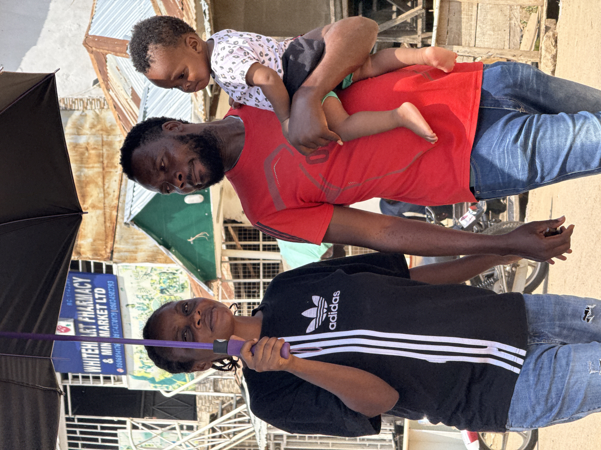 A family walks under an umbrella on a street in Abuja, Nigeria. A woman on the left holds the purple umbrella, shielding herself and the man next to her. The man on the right holds a young child, who is facing away from the camera. The woman wears a black t-shirt with white stripes and an Adidas logo, and blue jeans. The man wears a red t-shirt and blue jeans. The child wears a white t-shirt with a leopard print pattern and black shorts. In the background, there are buildings with corrugated metal roofs, a pharmacy sign that reads "WHITEHEART PHARMACY & MIN MARKET LTD," and a motorcycle. The ground is dirt and appears to be dusty.