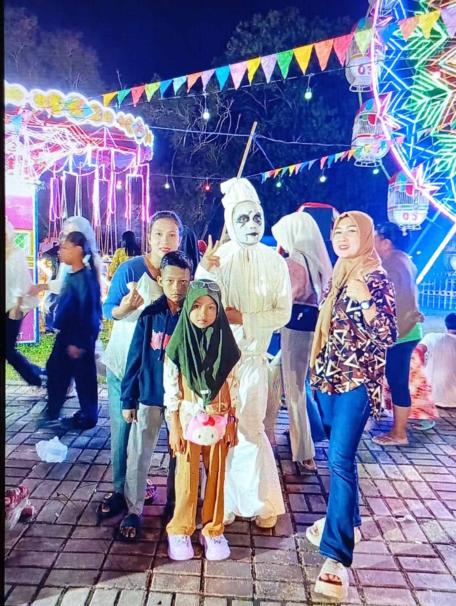 The image captures a nighttime scene at an outdoor event, possibly a carnival or festival, in Lhokseumawe, Indonesia. In the foreground, a group of people are posing for a photo. To the left, a person in a white costume with face paint stands with their arms out. Next to them, a woman in a blue and white shirt smiles at the camera, giving a thumbs-up. In front of her are two children; a boy in a dark hooded sweatshirt and a younger girl wearing a green hijab and a pink Hello Kitty bag. To the right of the group, a woman in a tan hijab and a patterned blouse smiles. The background is illuminated by vibrant lights from a Ferris wheel and other attractions, with colorful pennant banners strung overhead. The ground is paved with interlocking bricks.