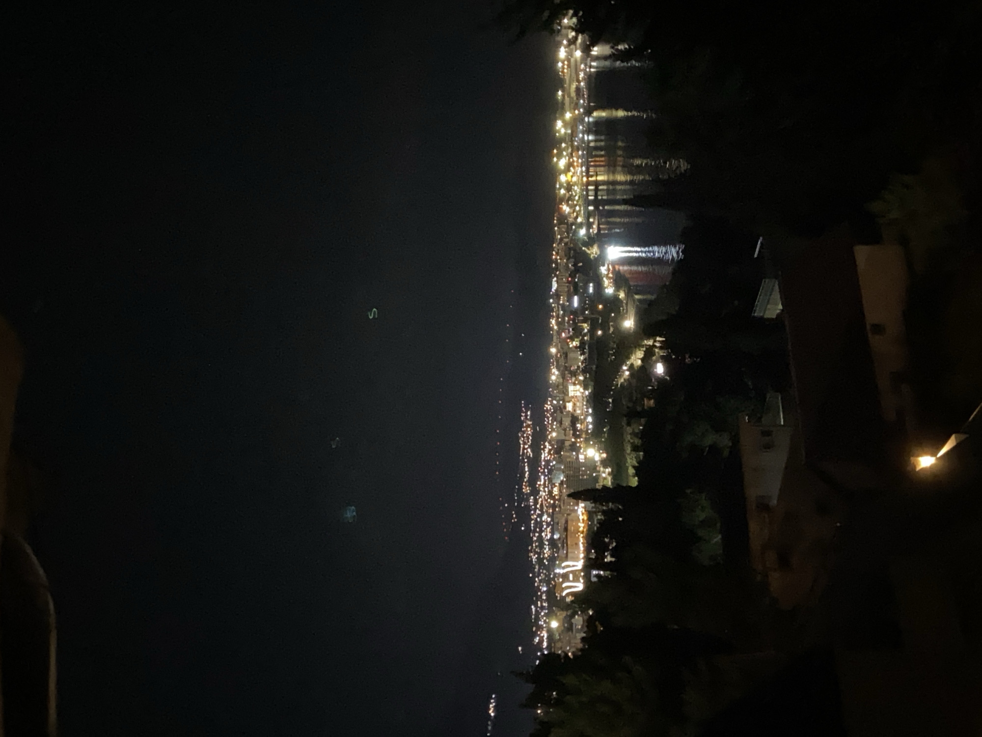 This is a night shot overlooking the city of Šušanj, Montenegro. The scene captures a coastal city illuminated by a multitude of lights, reflecting on the dark water of the bay. In the foreground, silhouettes of trees and buildings are visible, creating a stark contrast with the brightly lit cityscape in the background. The sky is dark, with a few faint, unidentifiable luminous objects scattered across it. The overall impression is one of a quiet, serene evening in a coastal town.