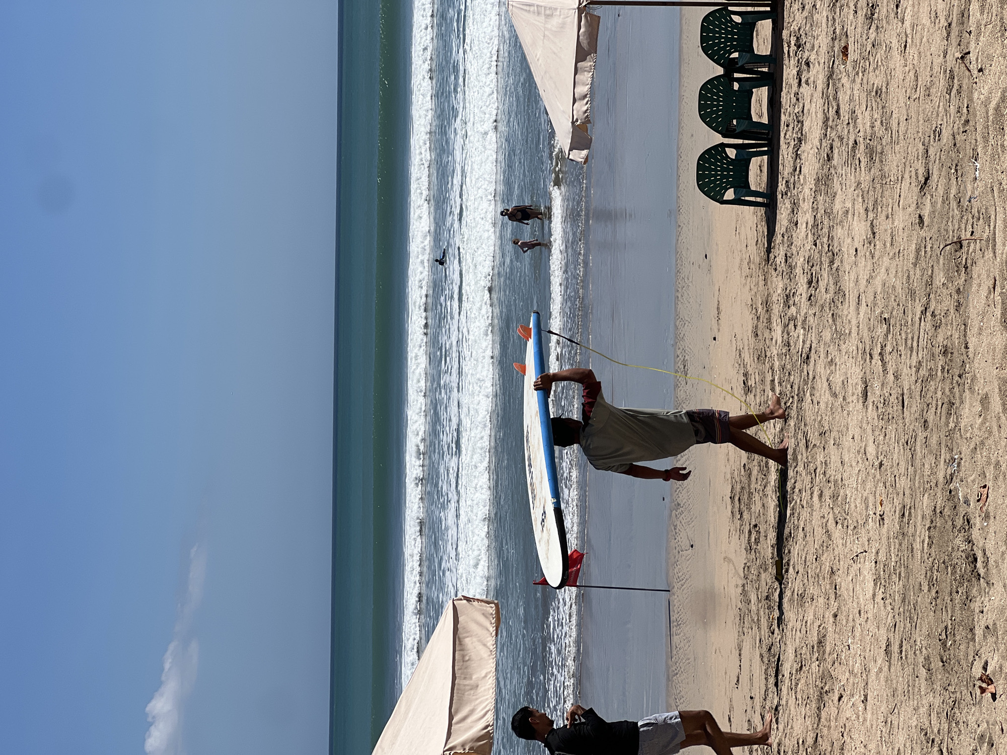 The image depicts a sunny day on Kuta Beach in Bali, Indonesia. In the foreground, two men are on the sand. One man, on the left, is wearing a black long-sleeved shirt and striped shorts. The other man, in the center, is carrying a surfboard over his head and is wearing a t-shirt and patterned shorts. He is walking towards the right. Behind them, the ocean has gentle waves, and several people are in the water. Some are standing, while one person is on a surfboard further out. Two beach umbrellas, one on the left and one on the right, provide shade. Three green plastic chairs are set up on the sand to the right, beneath the umbrella. The sky is clear blue with a few scattered clouds. A small red flag is planted in the sand between the two men. The sand appears wet closer to the water and dry further up the beach, with some debris visible on the surface.