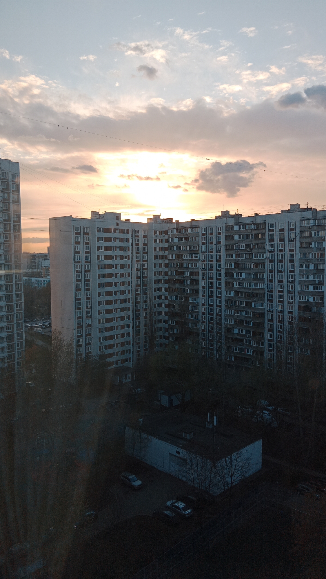 The image depicts a view of a residential area in Moscow, Russia, during a sunset. Several high-rise apartment buildings dominate the skyline. The buildings are characterized by their repetitive window patterns and grey concrete facades. In the foreground, there is a lower, possibly non-residential building with parked cars around it. Trees, mostly bare, are scattered around the buildings. The sky is filled with clouds tinged with orange and pink hues from the setting sun. Some birds are flying across the sky in the background. The photograph is taken from an elevated position, likely from within another building.