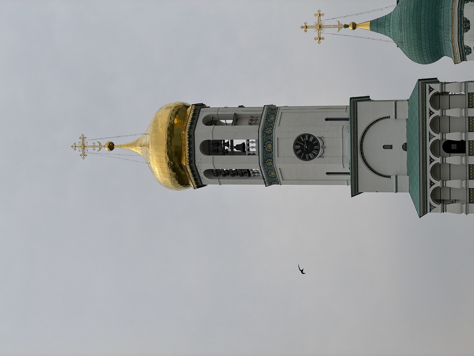 The image shows the Voskresensky New Jerusalem Monastery in Istra, Russia, on a cloudy day. The central focus is a white bell tower with a golden dome topped by a cross. The bell tower features several arches, decorative trim, a clock with Roman numerals, and a set of bells visible within the open arches. To the right, another green dome topped with a golden cross is partially visible. In the sky to the left of the bell tower, a small bird is captured in flight. The overall scene conveys a sense of architectural grandeur against a muted, overcast sky.