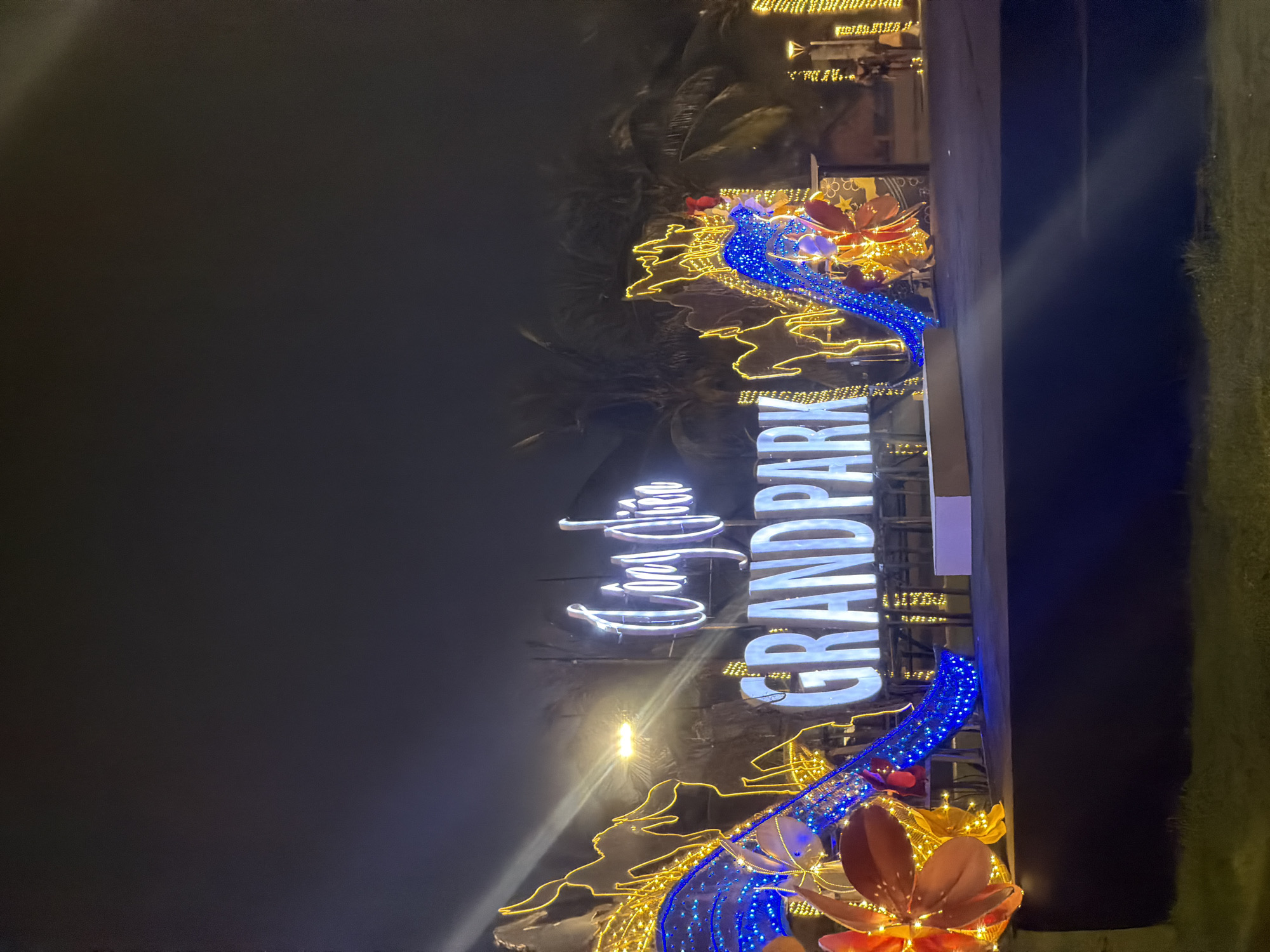The image shows a nighttime scene at Grand Park, located in Thu Duc City, Ho Chi Minh City, Vietnam. A large, illuminated sign with "GRAND PARK" in prominent white letters is the central focus. Above it, a smaller, cursive Vietnamese sign is also lit. The sign is surrounded by a festive and colorful display of lights.
On the left side of the sign, there are large, sculpted floral decorations with orange and red petals, intertwined with bright yellow and blue string lights. The blue lights appear to be shaped like flowing water or ribbons. There are also abstract, illuminated yellow outlines that resemble rock formations or organic shapes.
To the right of the main sign, the illumination continues with what appear to be strings of yellow lights decorating palm trees or other foliage. In the background, several people are visible, including one person standing near a lamppost and another appearing to be on horseback. The ground in front of the display is a dark, elevated platform, and beyond that, a grassy area is visible. The overall atmosphere is festive and vibrant, suggesting an event or a decorated public space. The sky is dark, indicating it is nighttime.