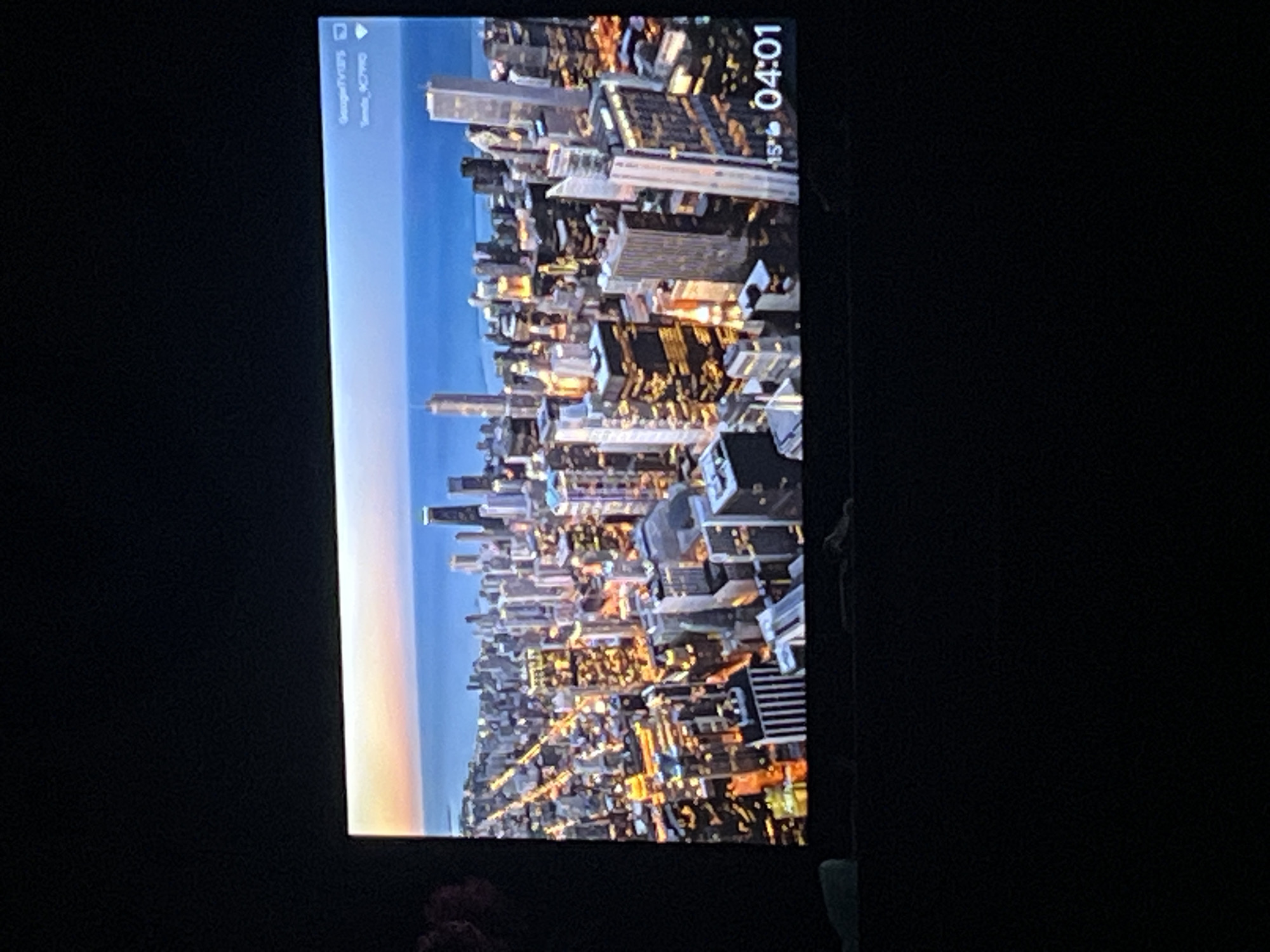 The image displays a wide shot of a city skyline at dusk, with numerous skyscrapers illuminated by interior lights. The city is situated on the coast of a large body of water, likely a lake or ocean, under a sky with a gradient of orange, pink, and blue hues. On the lower right corner of the screen, there is a time display showing "04:01" and a temperature reading of "15°". In the upper right corner, there are two lines of text; the top one reads "GoogleTV" and the bottom one appears to be a network identifier. There is no discernible connection to Zoriane, Ukraine.