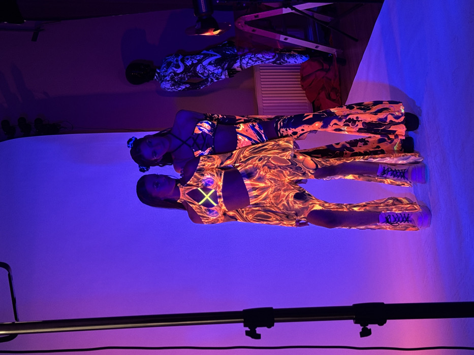 The image depicts an indoor photography studio located in Sofia, Bulgaria, illuminated primarily by ultraviolet light, casting a pronounced blue and purple hue across the scene. Two female models are positioned centrally against a seamless white cyclorama backdrop. The model on the left wears a long-sleeved crop top with a high collar and cut-out shoulders, featuring a neon yellow 'X' design on the chest. She is dressed in matching wide-leg pants with vertical slits along the sides. Her ensemble exhibits an orange and yellow "lava lamp" or "fire" pattern, fluorescing brightly under the UV light. She wears light-colored platform boots. The model on the right wears a two-piece outfit consisting of a strappy, laced-up bralette and matching wide-leg pants, also featuring a fluorescent orange, yellow, and black "lava lamp" pattern. Her hair is styled in two buns, each with a glowing accent. Both models display reflective or glitter embellishments around their eyes. To the right, a third figure, appearing to be a mannequin or person, stands in a full-body suit with a black and white abstract pattern that also reacts to the UV light. Visible studio equipment includes a black vertical stand in the foreground on the left, a white radiator on the far right wall, and a metallic A-frame ladder or stand to the far right. The floor is covered with a white sweep, extending from the backdrop.