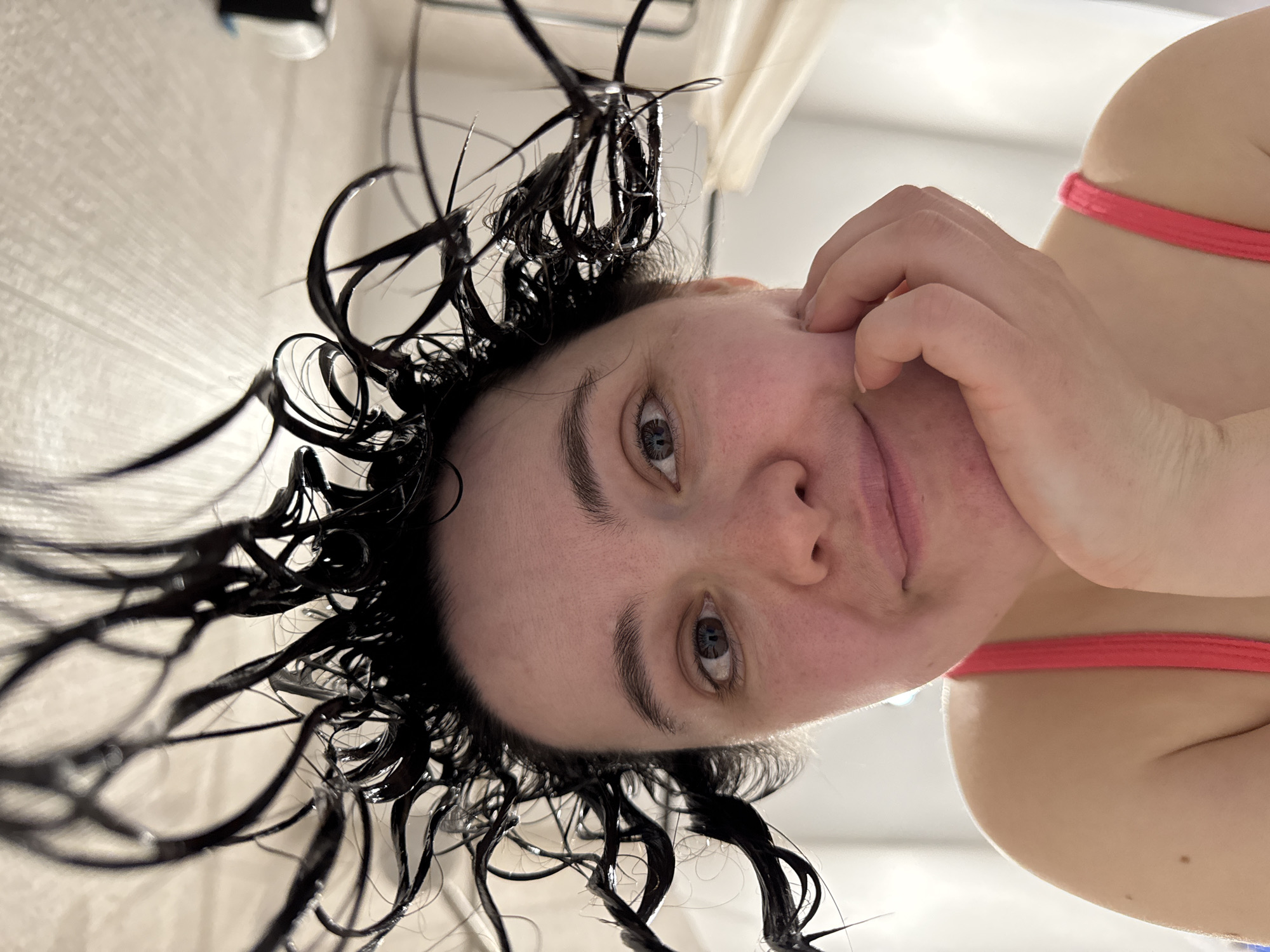 A selfie of a woman with wet, dark, curly hair. Her hand is touching her cheek. She has fair skin and light-colored eyes. A pink strap is visible near her shoulder. The background appears to be a bathroom with tiled walls. There is no indication of Izhevsk, Russia.
