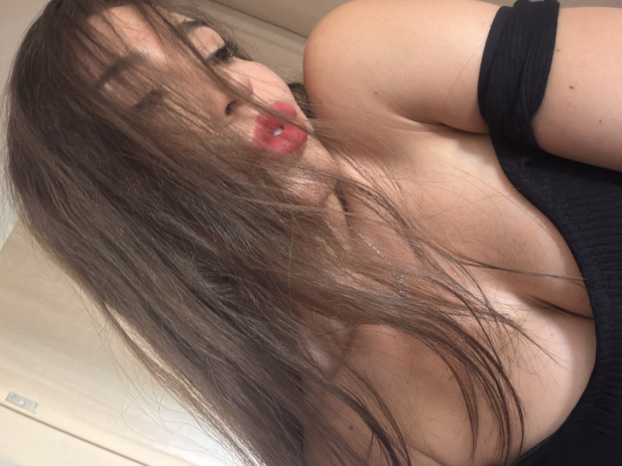 A woman with long brown hair covering her face is shown from the chest up. She is wearing a black off-the-shoulder top and has bright red lipstick on. A silver necklace is visible around her neck. The background appears to be an indoor setting with a light-colored wall and a door frame. The image was taken in Tersefanou, Cyprus.