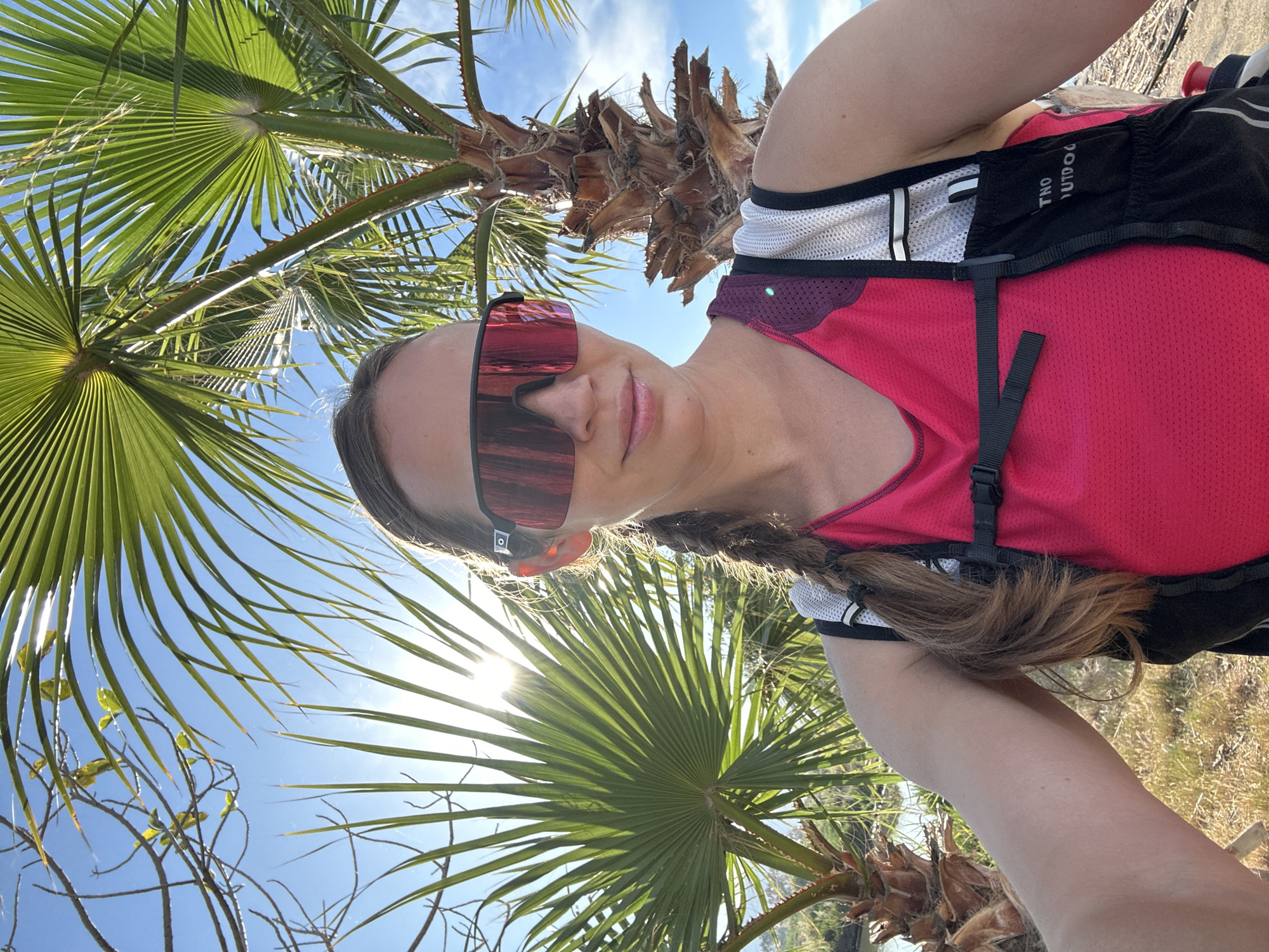 A selfie captures a fair-skinned woman with sunglasses, a braid, and a hydration pack in a sunny outdoor setting. She is wearing a pink and black athletic top. Palm trees dominate the background, with a clear blue sky visible through the leaves. The ground appears to be covered with dry grass. Based on the image's description, the location is likely in or near Fethiye, Turkey, although specific landmarks are not discernible.
