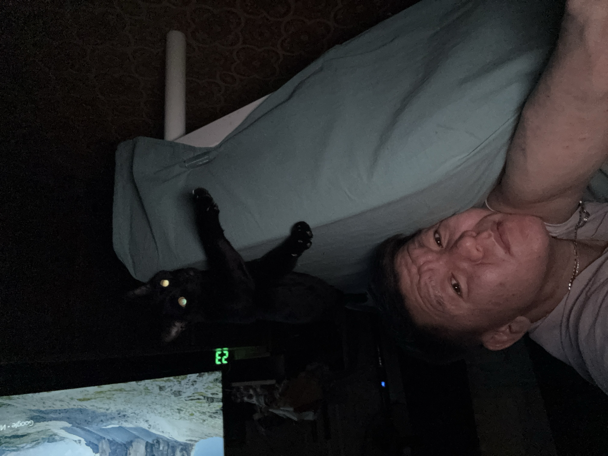 A dimly lit indoor scene shows a person and a black cat. The person, seen from the neck up, is facing the camera and slightly angled to the right. Their face is illuminated enough to discern features. A silver chain necklace is visible. Adjacent to them is a pale blue pillow. To the left of the pillow, a black cat with reflective yellow eyes lies in shadow. The cat's paws are visible. Below the cat is a monitor displaying a landscape. The number "23" is illuminated on a device near the monitor. A white pipe is visible in the background. The location is not evident in the image.