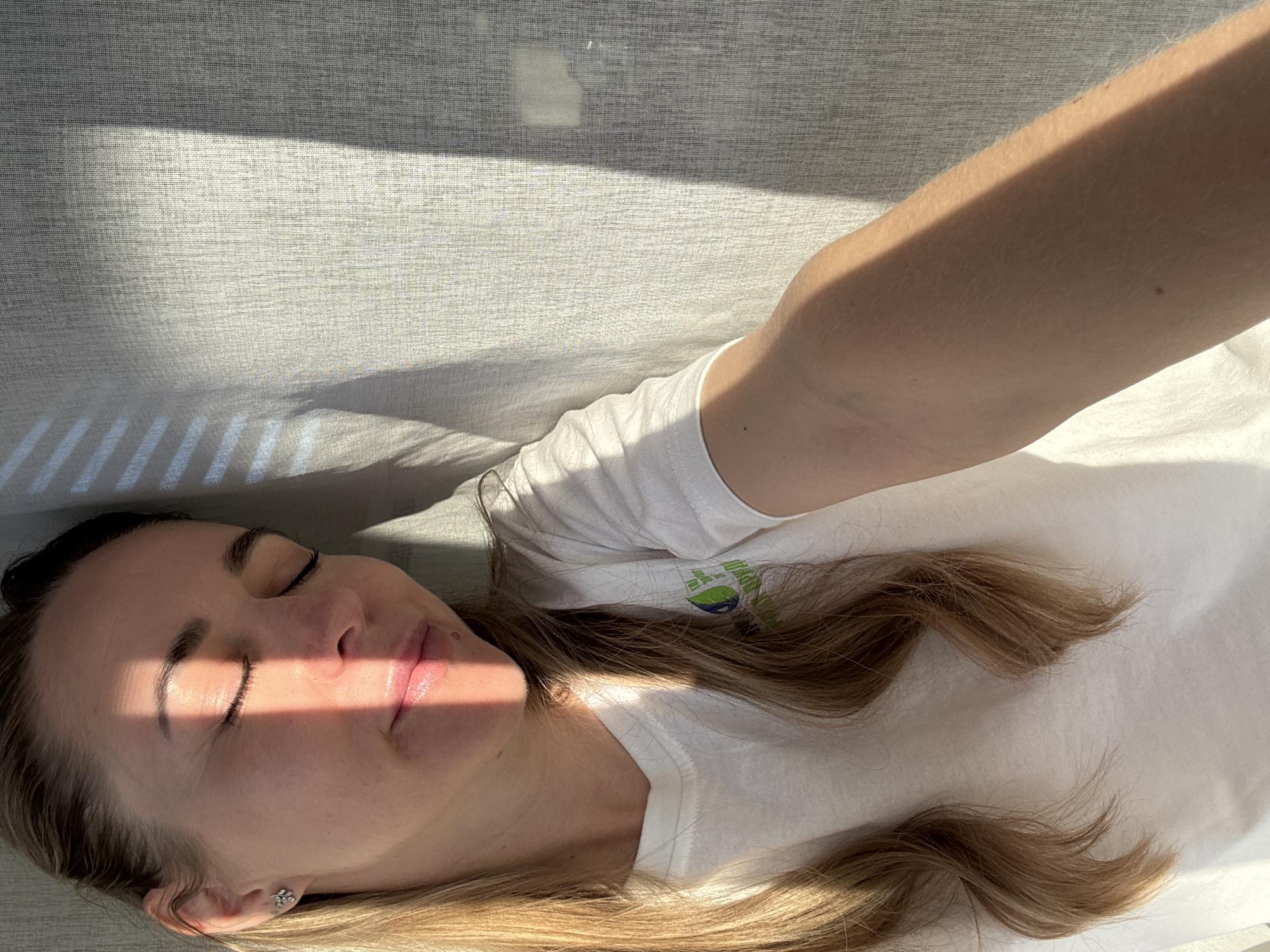 A woman lies in repose. Her eyes are closed. The sun illuminates her face, and her hair is tousled about her shoulders. She is wearing a white t-shirt. The location is undetermined. There is no evidence of the city of Chaikovsky, Russia.