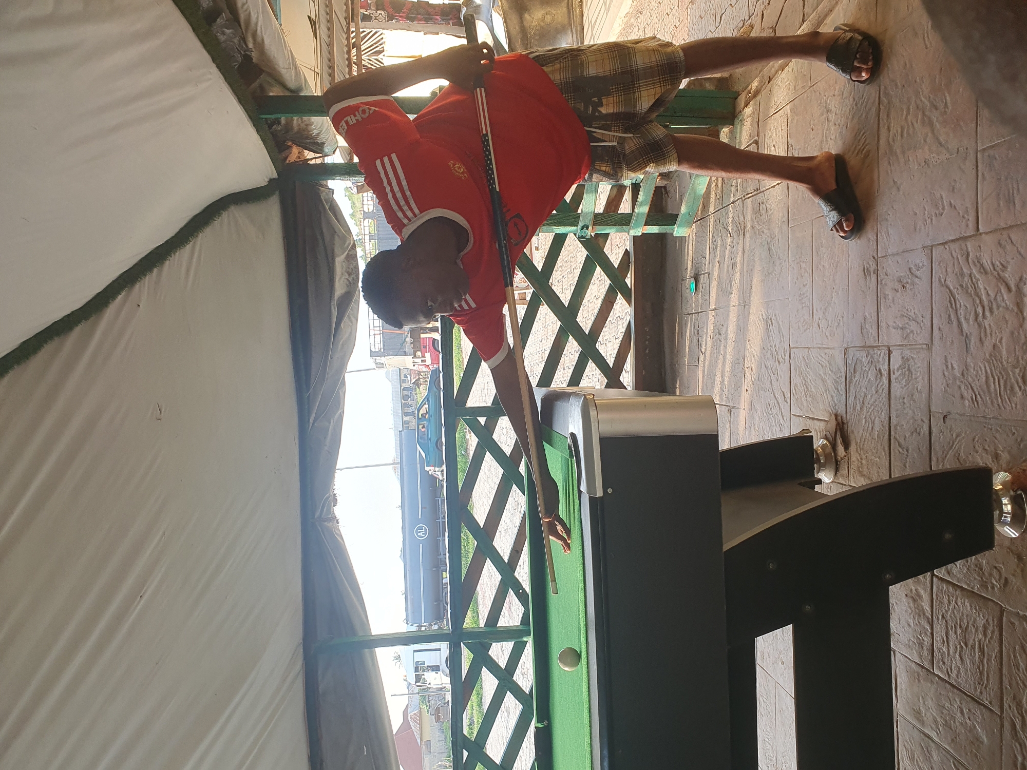 A young man is playing billiards on a pool table. He is wearing a red Manchester United t-shirt and plaid shorts. He is holding a pool cue and is about to strike a ball on the green felt surface. The pool table is black with a silver trim. The setting appears to be outdoors, under a covered area with a green wooden fence. In the background, there are some buildings and what looks like a train car. The time of day appears to be daytime, with sunlight casting shadows. The man's posture suggests concentration as he plays the game. The overall mood is casual and relaxed, typical of a recreational activity. The image seems to be taken in Lokoja, Nigeria, as per the location context.