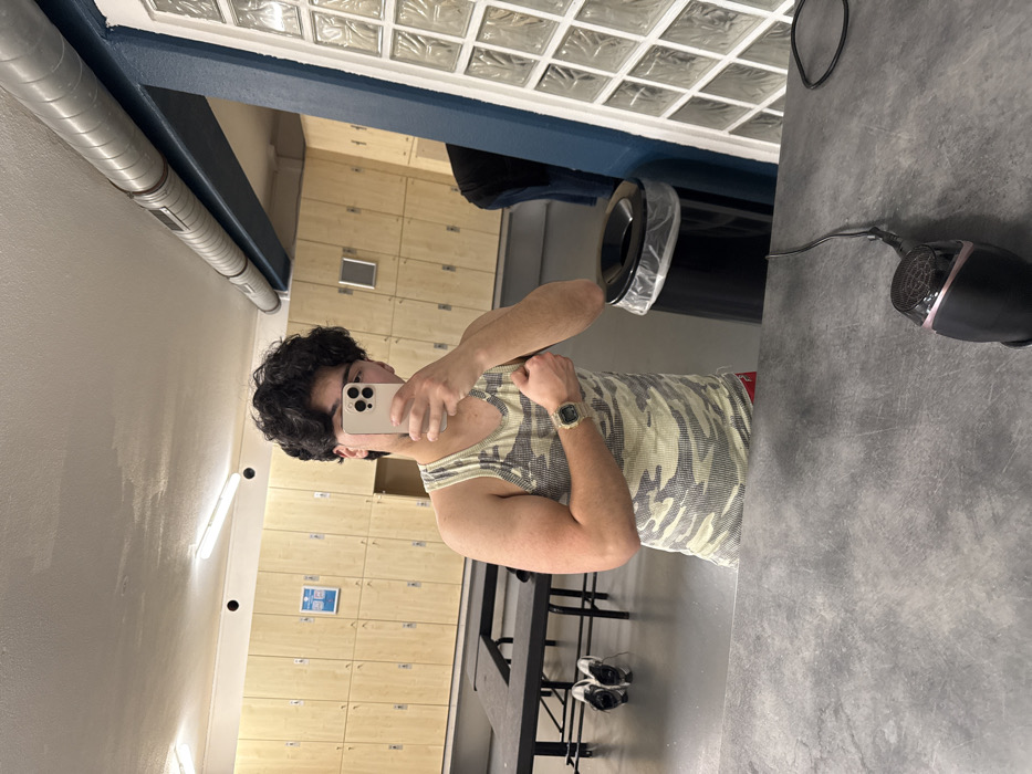 The image displays an indoor scene within a locker room or changing area, specified as being located in Geispolsheim, France. A male individual is reflected in a large mirror, appearing in the center of the frame. He possesses dark, curly hair and is wearing a light camouflage-patterned ribbed tank top with red shorts visible at the waist. His face is obscured by a light-colored smartphone, which he holds in his left hand, angled to capture a mirror selfie. His right arm is bent, flexing the bicep, and a digital wristwatch is visible on his left wrist.
In the immediate foreground, a dark gray countertop features a black and pink hairdryer with its power cord, resting flat. The background, visible through the mirror's reflection, consists of a long wall lined with numerous light wood lockers, each with a blue label holder. Two elongated, black benches are positioned in the mid-ground, with a pair of black and white athletic shoes resting on the floor beneath one. To the right, a black cylindrical trash can with a white plastic liner is present. The ceiling above the lockers shows fluorescent tube lighting, a black circular vent, and exposed silver-gray ventilation ducts running parallel to a blue structural beam. A section of a white, textured wall, possibly composed of glass blocks, is visible on the far right, next to a blue wall portion.