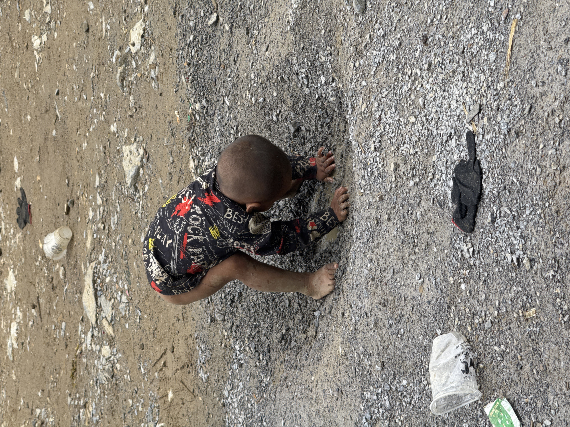 A young, bare-footed child in a black shirt with red and white printed designs is on the ground, hands outstretched, in Phố Bảng Commune, Vietnam. The ground is a mix of dirt, gravel, and various debris. A black object (possibly a glove) and a white plastic cup are on the ground near the child.