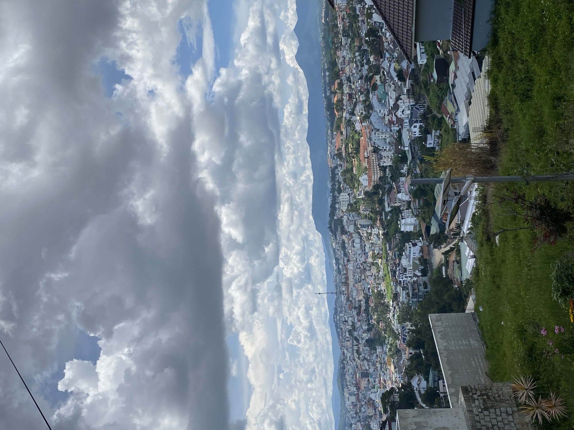An aerial, slightly angled, daylight, medium-shot captures a panoramic view of the city of Da Lat, Vietnam. The urban sprawl, characterized by numerous buildings with red-tiled roofs and white walls, covers a vast expanse of rolling hills that recede into a hazy, mountainous backdrop under a partially cloudy sky. The foreground is dominated by a steep, grassy incline with a rough, concrete wall and brick structure on the left, hinting at ongoing construction or a viewpoint. A lamppost with solar panels stands prominently in the middle of the grassy area. The sky is a dramatic mix of dark gray and bright white clouds with patches of blue visible, suggesting a dynamic weather system. The overall impression is one of a densely populated, picturesque city nestled within a lush, natural landscape.