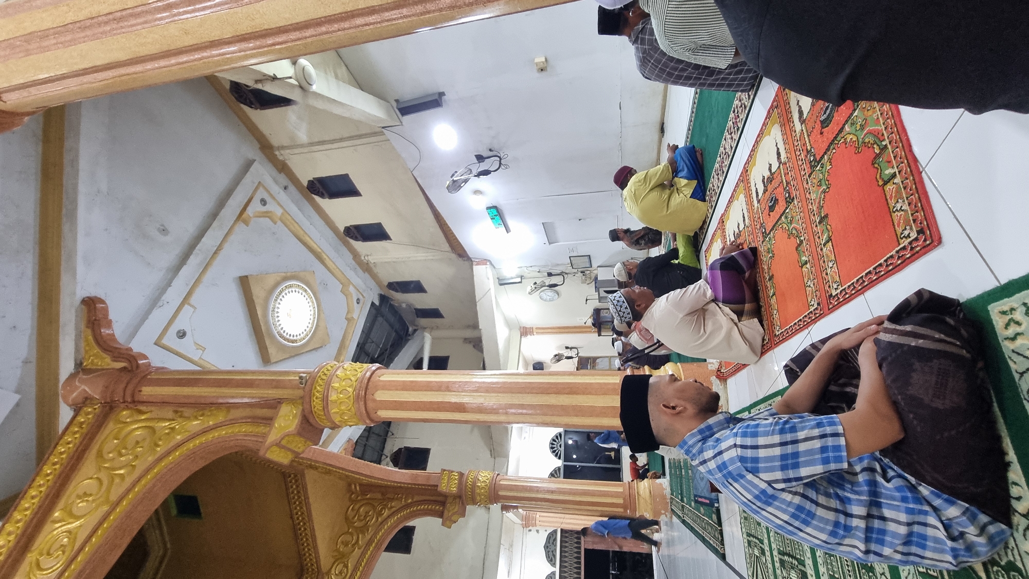 The image depicts the interior of a mosque in Manetunong, Indonesia, where several men are seated on prayer mats. The architecture features ornate columns with golden and pink detailing, supporting a ceiling with decorative panels and lighting. The men, dressed in traditional Islamic attire, are facing forward, presumably in prayer or contemplation. Some are wearing kufis, while others have their heads uncovered. The floor is covered with green prayer mats, with colorful patterned mats placed in front of some individuals. The environment appears to be an indoor prayer hall, with a white wall in the background and windows visible. A digital clock is mounted on the wall. The lighting is a mix of overhead lights and ambient light from windows. The perspective is from a low angle, focusing on the seated individuals and the surrounding architecture.