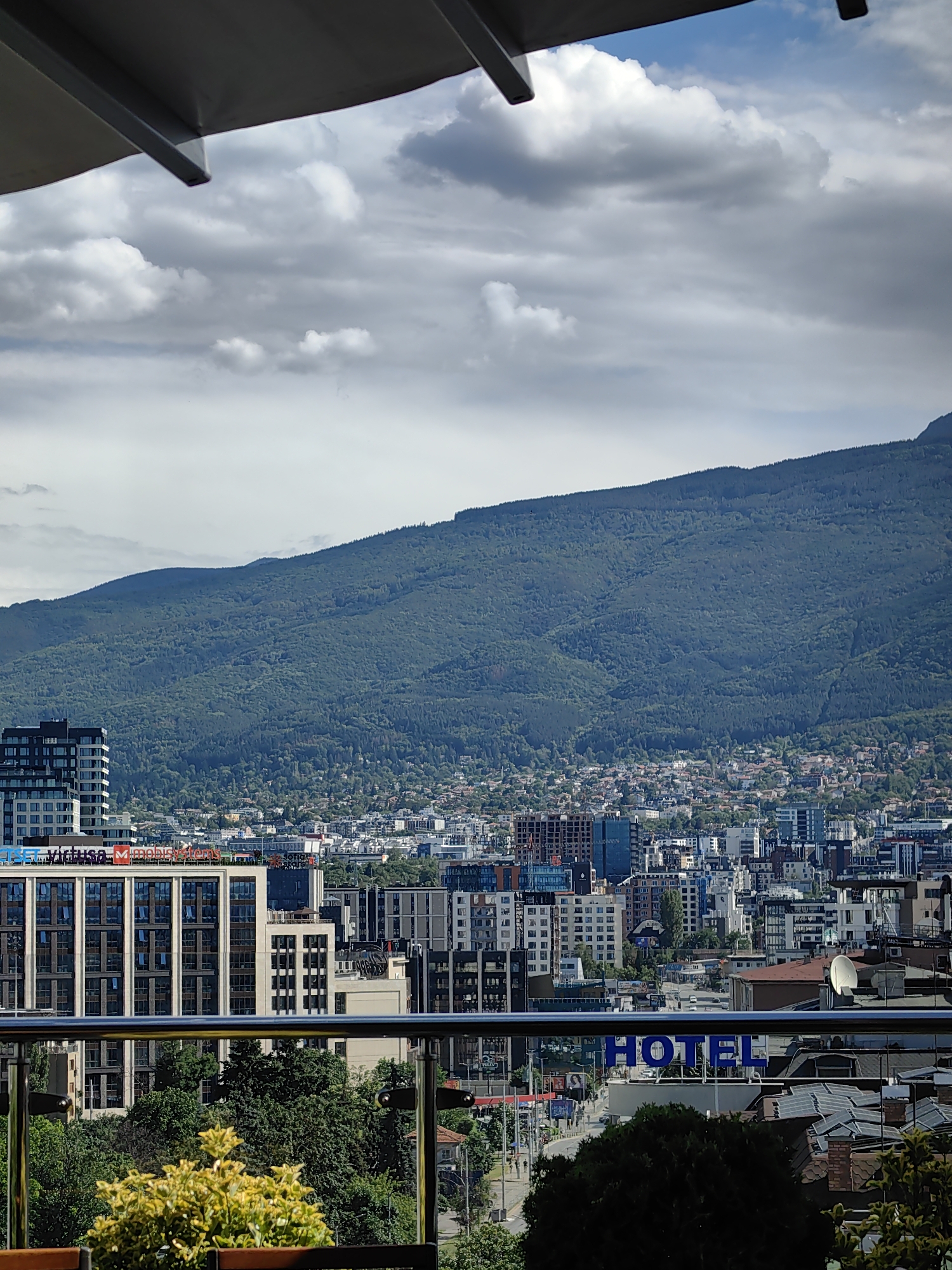 The image is a telephoto shot of the city of Sofia, Bulgaria, with a mountain range in the background. In the foreground, there is a balcony with a metal railing and a yellow bush. Beyond the balcony, the cityscape is visible, featuring various buildings of different heights and architectural styles. Notably, a tall, modern building with a glass facade stands out. In the mid-ground, there are more buildings, some appearing to be office buildings, and a street with some traffic can be seen. A prominent sign with the word "HOTEL" in large blue letters is visible on one of the buildings. The background is dominated by a large, densely forested mountain, which forms a significant backdrop to the urban landscape. The sky above is partly cloudy, with some bright white clouds against a blue sky. The overall impression is a panoramic view of a city situated at the foot of a mountain.