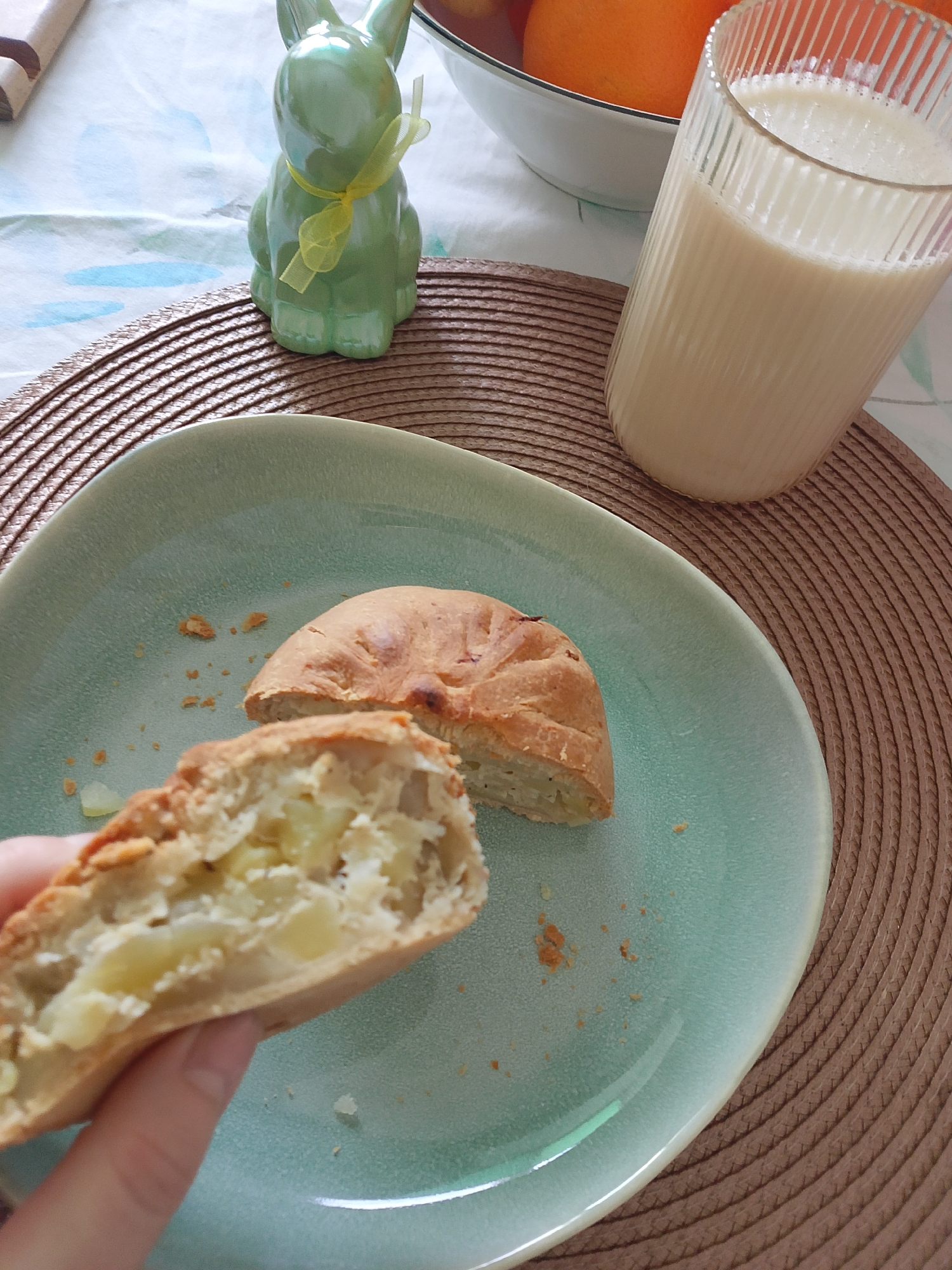 This image depicts a tabletop scene, reportedly located in Izhevsk, Russia. The surface is covered by a light-colored tablecloth featuring faint blue patterns, beneath a circular woven brown placemat. A light teal-green plate with an irregular rim occupies the central foreground. On the plate, two halves of a baked pastry, characterized by a golden-brown crust and a chunky, pale-yellow filling, are present. A human left hand holds one half of the pastry, exposing its interior. Minor pastry crumbs are scattered on the plate. To the right of the plate, a clear, ribbed glass contains an opaque, light-beige liquid, consistent with a dairy or plant-based milk. In the background, a white bowl with a dark rim contains several whole orange fruits. Adjacent to the bowl, a green ceramic rabbit figurine with a yellow ribbon tied around its neck stands upright. A segment of a light-colored wooden object is partially visible in the upper-left quadrant.