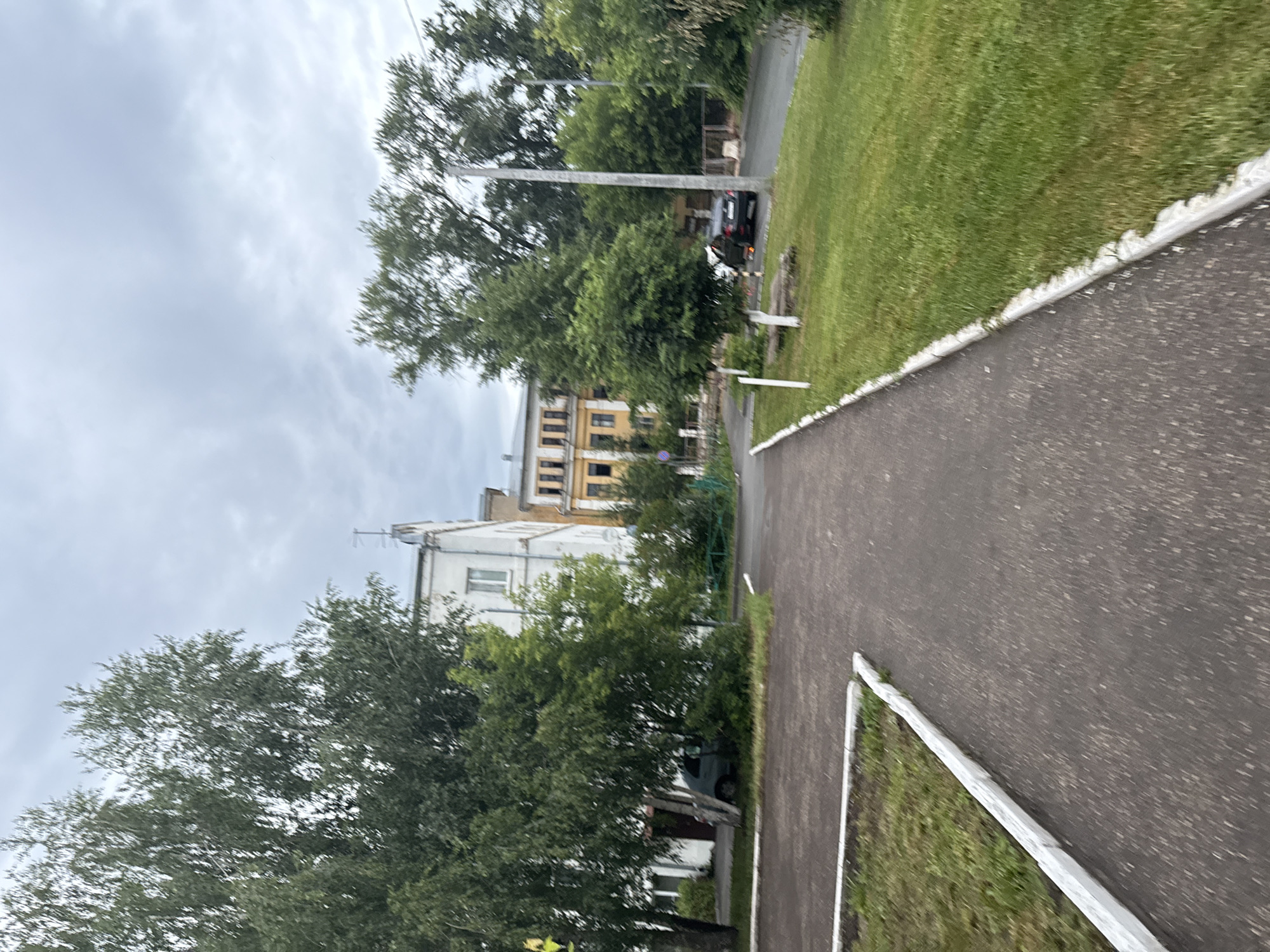 The image shows a view of a street with a paved walkway and a grassy area. Several trees line the walkway and the street. In the background, there are buildings, including a multi-story building with a yellow facade and another white building with windows. A black car is parked on the street. The sky is overcast, suggesting a cloudy day. The perspective suggests the photo was taken from a moving vehicle or while walking along the path. The scene appears to be a residential or public area in Kazan, Russia. There is no visible text in the image. The overall mood is neutral, capturing an ordinary urban or suburban landscape on a day with unfavorable weather conditions.