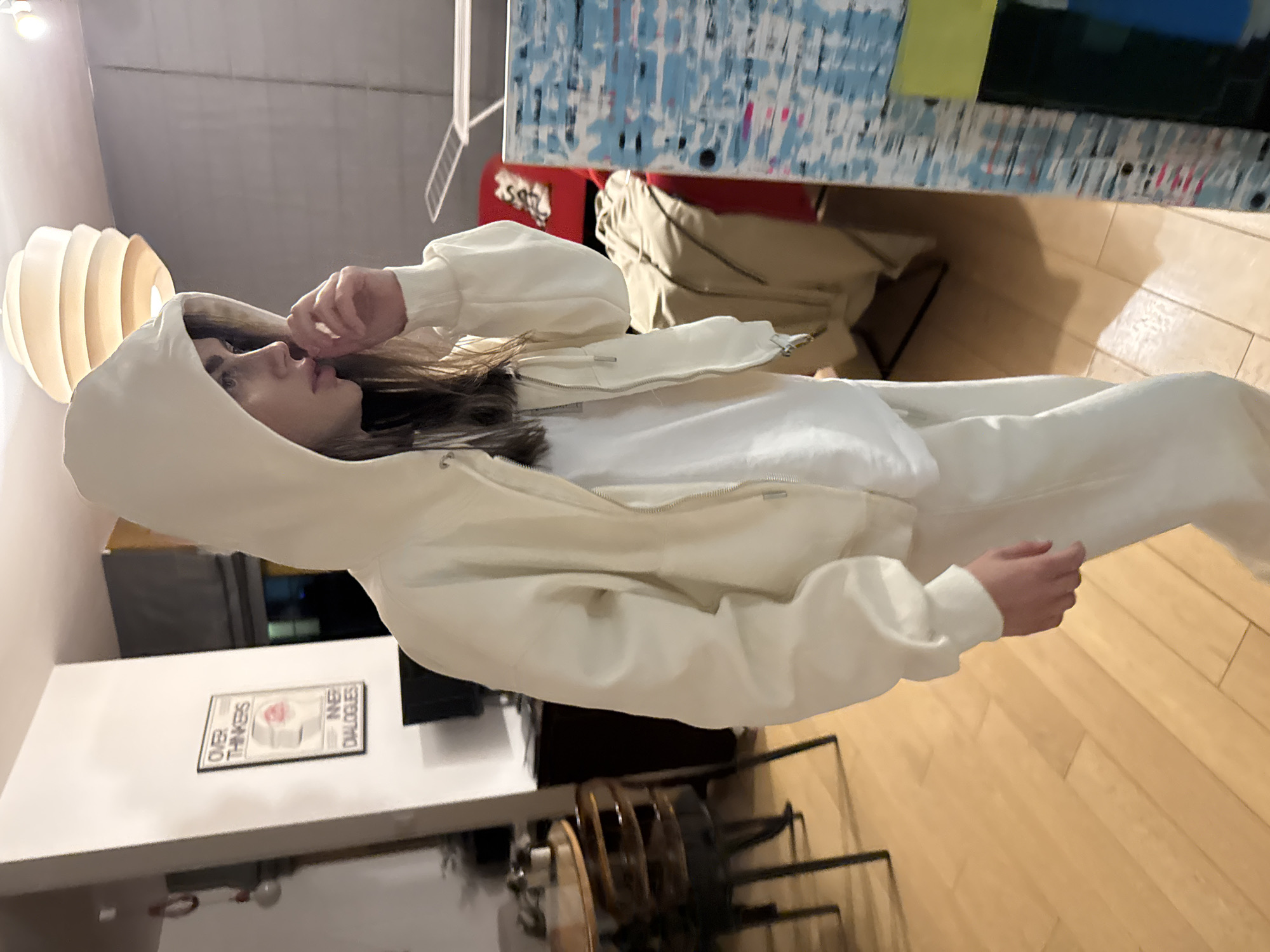 A person wearing a white hoodie and sweatpants is standing in an interior space. The space contains wooden floors, chairs with metal frames, and white walls with a framed poster titled "Over Thinkers Inner Dialogues." In the background, there is a lamp and a painting with blue and white colors. The specific location in Warsaw, Poland, cannot be determined based on the provided image details.