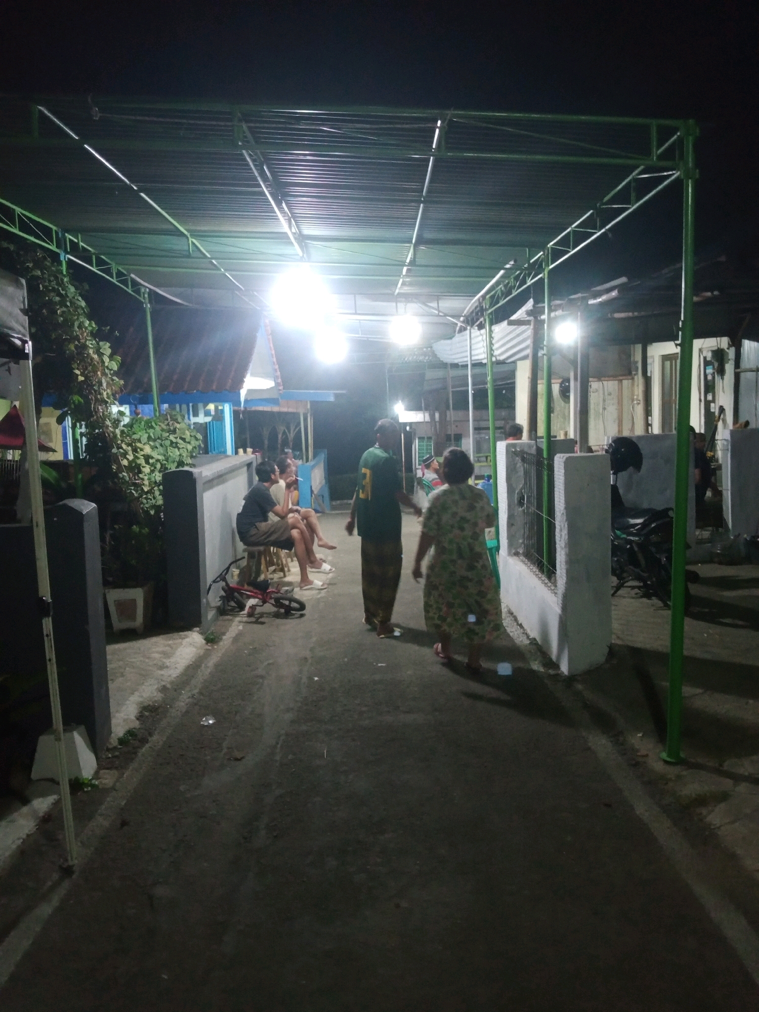 The image captures a nighttime scene in what appears to be a residential area in Rejasari, Indonesia, under a covered walkway illuminated by bright lights. In the foreground, two individuals are walking away from the camera. The person on the left is wearing a green shirt and patterned pants, while the person on the right is wearing a floral print dress. To the left, a few people are seated on stools by a low wall, with a small red bicycle lying on the ground nearby. In the background, the covered structure continues, with several buildings visible through the gaps. A motorcycle is parked near a white pillar. The environment is characterized by dark asphalt paving, low-lying vegetation, and a corrugated metal roof supported by green metal framing. The overall atmosphere suggests a casual gathering or evening activity within the community.