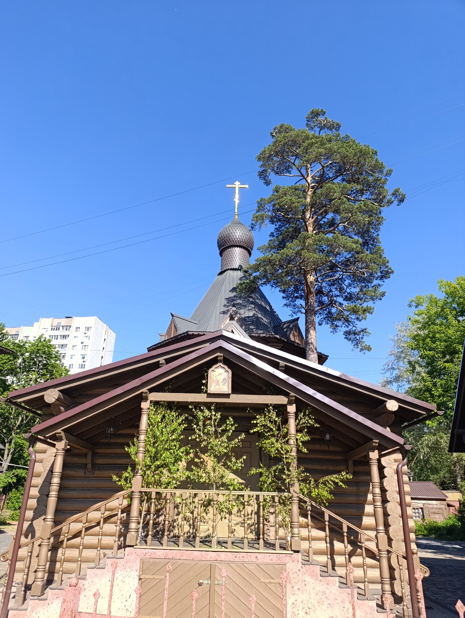 This image depicts the Church of the Kazan Icon of the Mother of God, located in the Vykhino-Zhulebino District of Moscow, Russia, specifically at 20 Saratovskaya Street.
The scene is dominated by a small Orthodox church constructed from horizontal light brown wooden logs, featuring a prominent dark gray onion-shaped dome topped with a golden Orthodox cross. The church has a steeply pitched dark gray shingled roof. Its main entrance is fronted by a covered porch with a dark brown trim roof and matching dark brown wooden railings and balusters. A set of faded reddish-pink stone or concrete steps leads up to this porch, with a low, similarly colored metal fence or gate visible at the bottom. Small green leafy plants grow on the porch, partially obscuring the front wall. An icon depicting religious figures is mounted on the gable of the porch roof, and a dark lantern hangs from the porch ceiling. A dark brown drainpipe runs down the left side of the building.
Behind and to the right of the church, a tall, mature pine tree with dark green needles stands prominently, its crown extending above the church dome. In the upper-left background, a multi-story, light-colored residential building with numerous windows is partially visible, indicating an urban or suburban setting. Several thin black utility lines traverse the clear, bright blue sky overhead. No people or animals are present in the frame. The scene is illuminated by strong sunlight, casting distinct shadows. A paved or cobbled path is visible on the far right.
